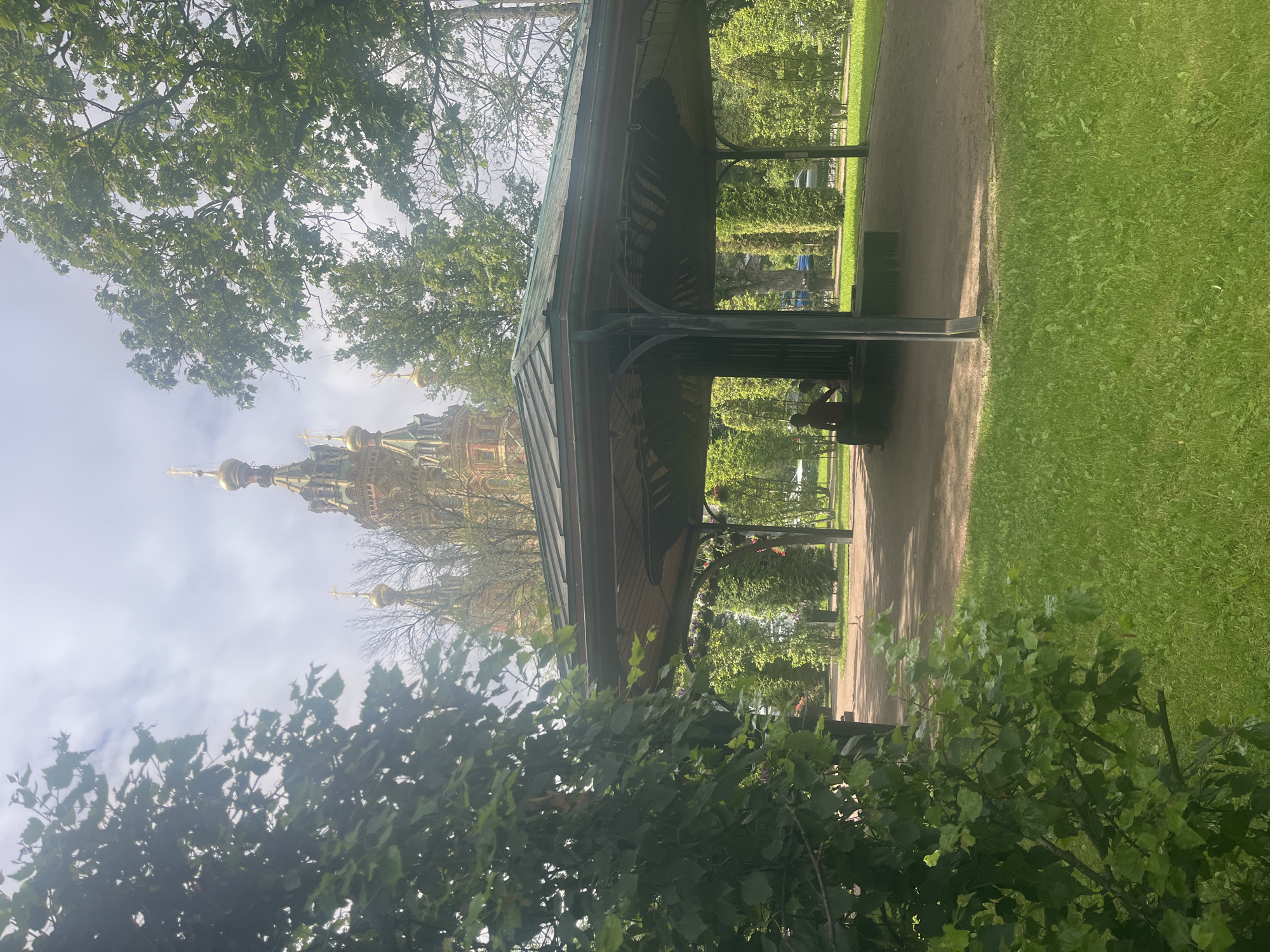 The image captures a sunny day in Peterhof, Russia, showcasing the grounds of the Peterhof Palace complex. In the foreground, lush green grass carpets a lawn that slopes gently towards a gravel pathway. A large, open-air pavilion with a dark, sloped roof and intricate metalwork supports dominates the middle ground. Two individuals, partially obscured by the pavilion's structure, are seated within its shaded interior.
Beyond the pavilion, a pathway winds through manicured trees and gardens. In the background, the distinctive ochre and copper domes and spires of the Peterhof Palace are visible, topped with golden crosses, peeking through the dense green foliage of mature trees. The sky above is a mix of blue with scattered white clouds, suggesting pleasant weather conditions. The overall scene depicts a serene park-like setting within a historically significant Russian landmark.