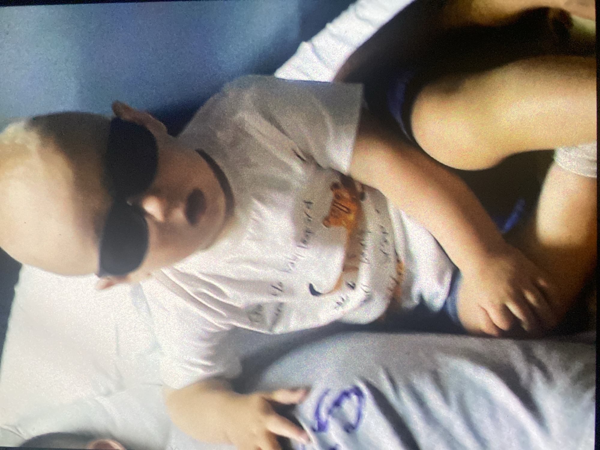 A child with blonde hair is lying on a bed wearing dark sunglasses and a white t-shirt with a graphic design. The child's mouth is slightly open. The bed has a light blue and white patterned sheet and pillow. The location appears to be indoors, but there are no clues to suggest the city of Saint Petersburg, Russia.