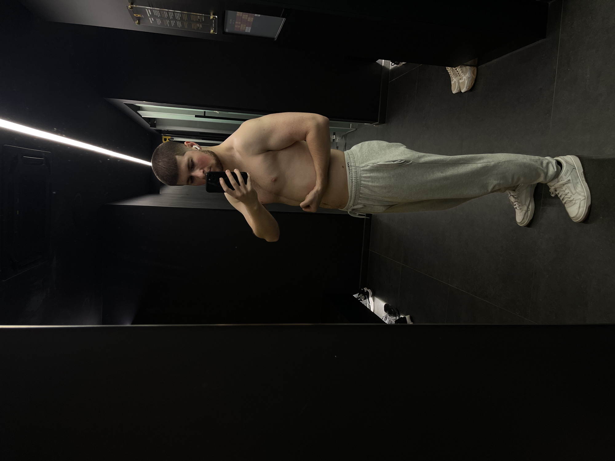 A shirtless man stands in a room with dark, possibly black, walls and floor, wearing grey sweatpants and white sneakers. He's holding a smartphone in his left hand, presumably taking a selfie. He has short hair, is wearing an earbud in his left ear, and is positioned slightly sideways to the camera. The scene appears to be a gym or dressing room. The presence of a sign on the wall and another pair of shoes on the floor suggests it could be a locker room setting. The location in Varna, Bulgaria cannot be confirmed from this image.