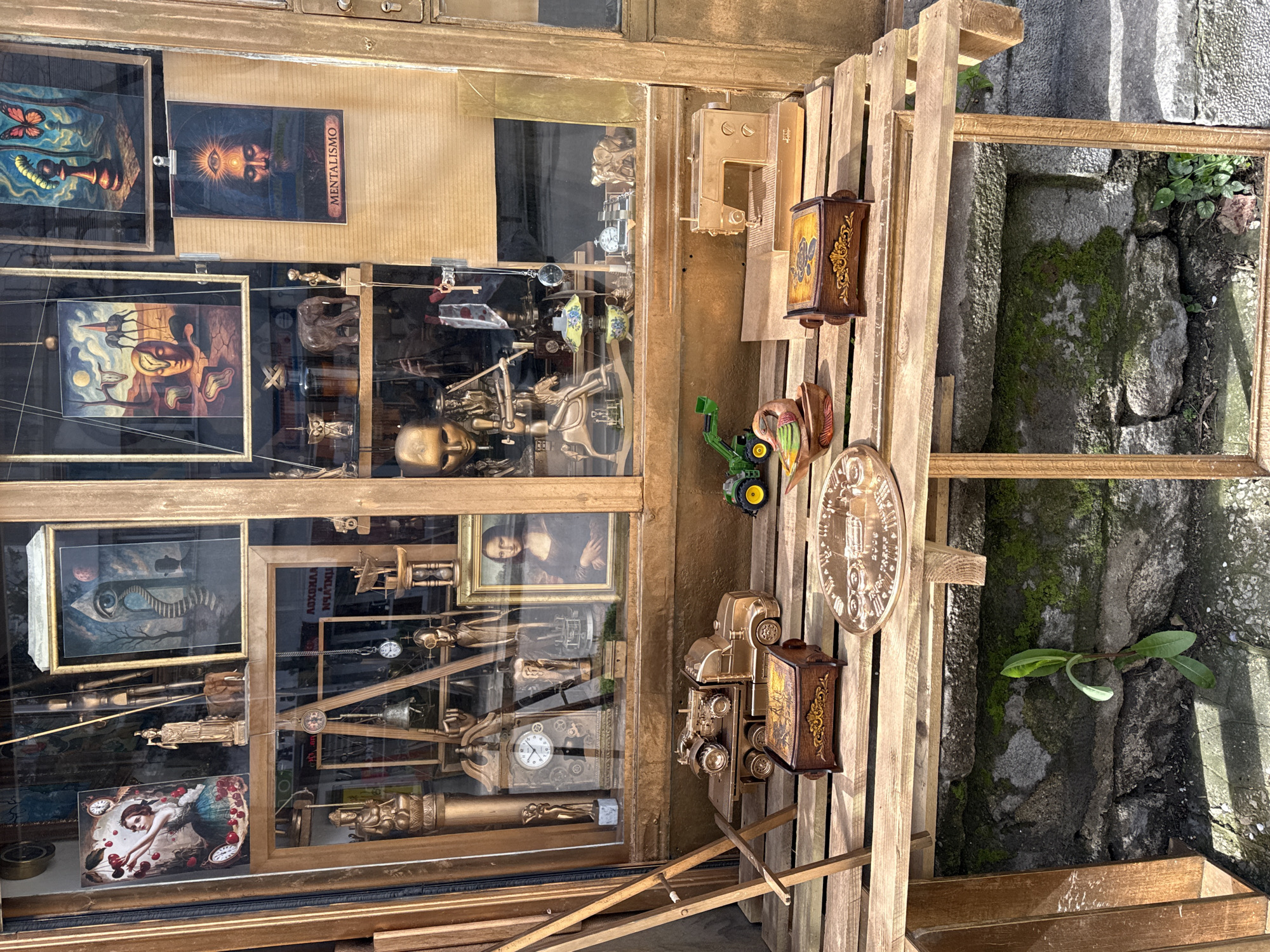The image shows a shop window display in Varna, Bulgaria. Inside the window, various framed artworks are visible, including pieces resembling Surrealist art, a portrait of a woman, and others with abstract themes. Below these, there are golden artifacts, including miniature statues, clocks, and decorative columns, all arranged in a complex, tiered display. To the right of the window is a wooden structure, possibly a pallet or display stand. Attached to this structure are additional objects, including decorative boxes, a small green tractor, and a round, ornate plaque. Behind the structure, a stone wall with moss growth and some vegetation is visible.