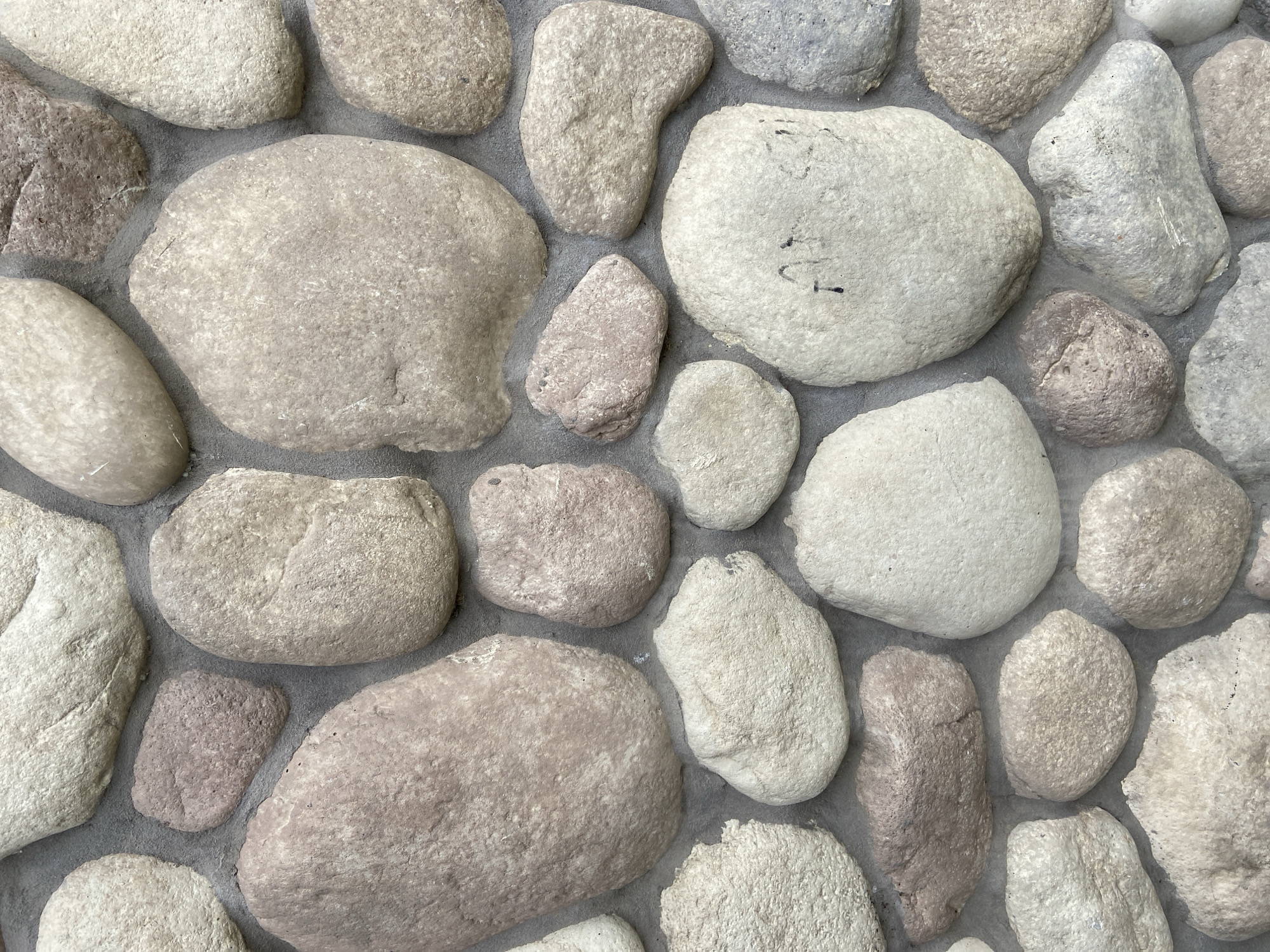 The image displays a close-up, detailed view of a textured wall constructed from irregularly shaped, smooth, rounded river stones of various sizes and colors, ranging from pale beige and cream to muted pinks and grays. The stones are set into a gray mortar that fills the gaps between them, creating a rustic and natural aesthetic. Some stones show subtle variations in their surface texture, with a few exhibiting faint markings or darker inclusions. The lighting appears to be natural daylight, casting soft shadows that accentuate the three-dimensional quality of the stonework. There are no people, animals, or discernible objects other than the stones and mortar. The specific location in Balashikha, Russia, cannot be determined from the visual information provided in the image itself.