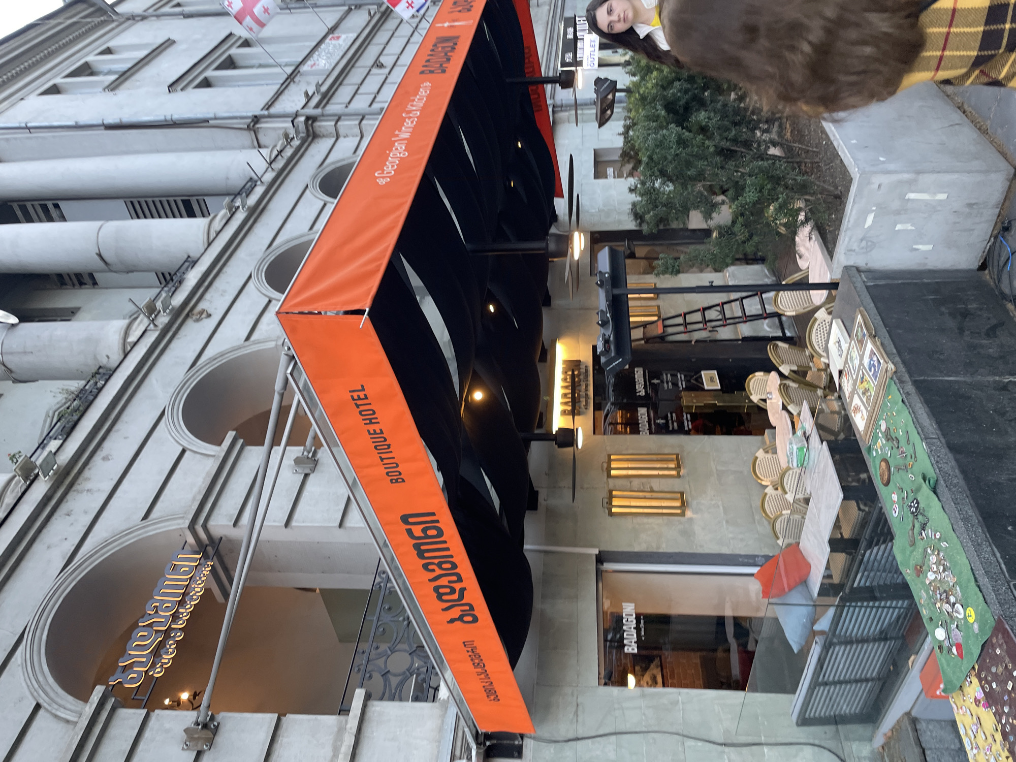 This image captures a vibrant street scene in Tbilisi, Georgia, focusing on the facade of a multi-story building featuring classical architectural elements like arched windows and pilasters. A Georgian flag proudly flies from the upper part of the building.
A large, bright orange awning extends over the sidewalk, advertising commercial establishments. Text on the awning, visible in both Georgian script and English, reads "BOUTIQUE HOTEL" and "Badagoni" (ბადაგონი), further clarified as "Georgian Wines & Kitchen" (ქართული ღვინოები & სამზარეულო). Below this, outdoor seating with light-colored wicker chairs and small tables suggests a cafe or restaurant setup. Another sign on the building in Georgian script appears to read "შალამან" (Shalaman).
On the right side of the image, a young woman with long, dark hair, wearing a light top and a yellow and black plaid jacket or shawl, looks towards the camera. The sidewalk area also features a grey concrete planter box with green foliage and a street vendor's display: a green cloth laid out with numerous small items for sale, including keys, badges, and brooches. Further back, another sign with "BADAGONI" is visible on a window, and an "OUTLET" sign is discernable in the distance.
The lighting suggests it's daytime, possibly overcast or late afternoon, as some artificial lights are on under the awning and inside the establishments. The scene depicts a lively urban environment, combining hospitality, retail, and local street vending.