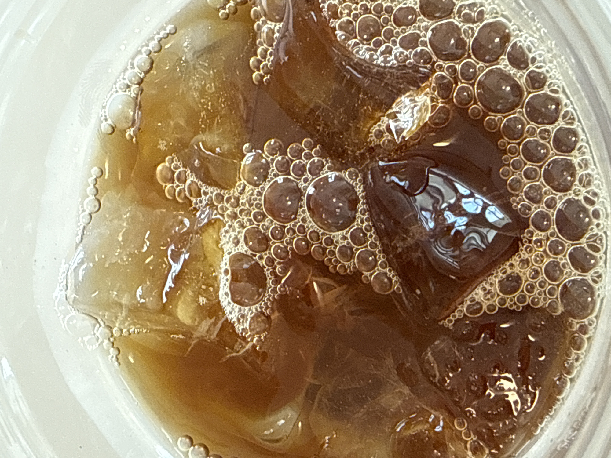 The image is a close-up, overhead shot of an iced coffee in a clear glass. The coffee is a dark brown color and is filled with ice cubes, some of which are partially submerged. The surface of the coffee is covered in a layer of small bubbles, indicating it has recently been stirred or poured. The lighting suggests it is daytime. There are no people or identifiable landmarks visible, and thus no specific location in Tel-Aviv, Israel can be determined from the image.