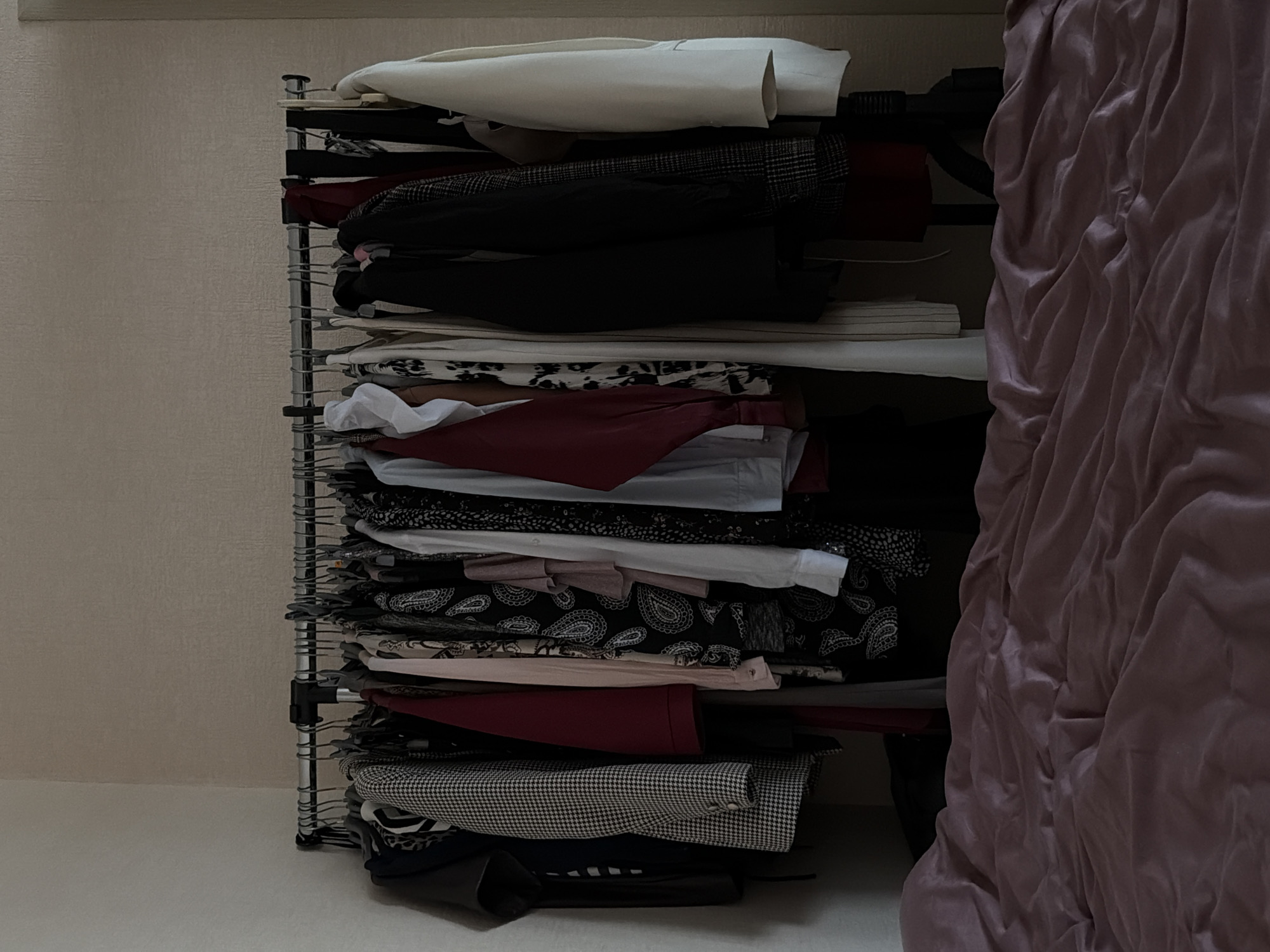 A disorganized, overloaded wire shelving unit is visible within what appears to be a closet. The shelves are stacked high with various garments, including shirts, blouses, and what appears to be a jacket with a houndstooth pattern. A dark, possibly black, item is visible at the bottom of the shelving unit, and more clothing items are piled up on the floor beneath it. To the right of the shelving unit is a wrinkled, rose-colored fabric, possibly a curtain or bedding, which adds to the crowded appearance. The setting has neutral wall and floor colors with no clear evidence of location in Izhevsk, Russia.