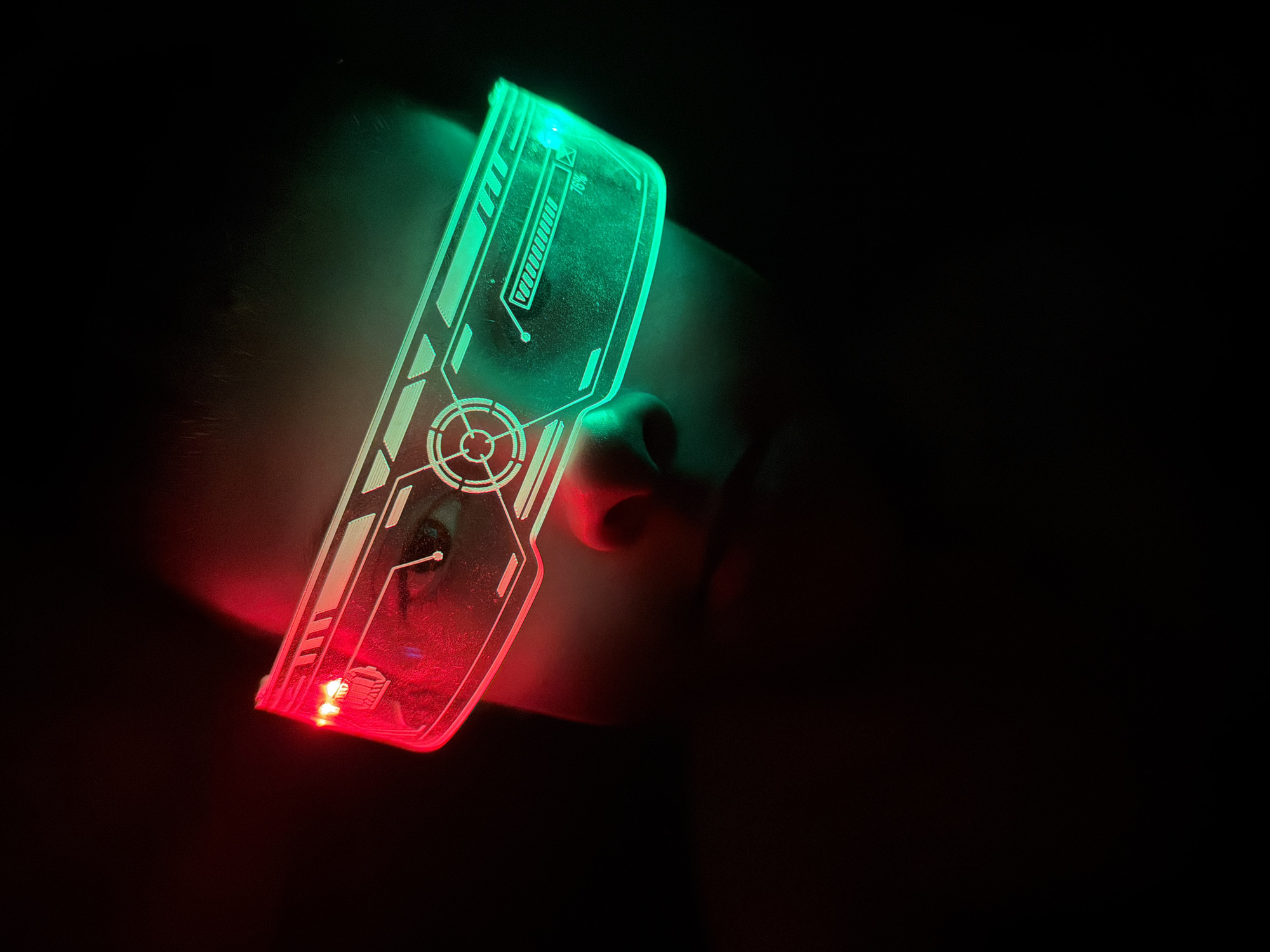A close-up, dimly lit image shows a person wearing futuristic red and green LED glasses. The glasses have a transparent frame with etched designs that glow, resembling a heads-up display (HUD). The upper part of the glasses emits a green light, while the lower part glows red. The person's face is partially visible behind the glasses, illuminated by the light they emit. The background is completely dark, contributing to the image's focus on the illuminated glasses and the person's face. There is no indication of the city of Votkinsk, Russia, in this image.