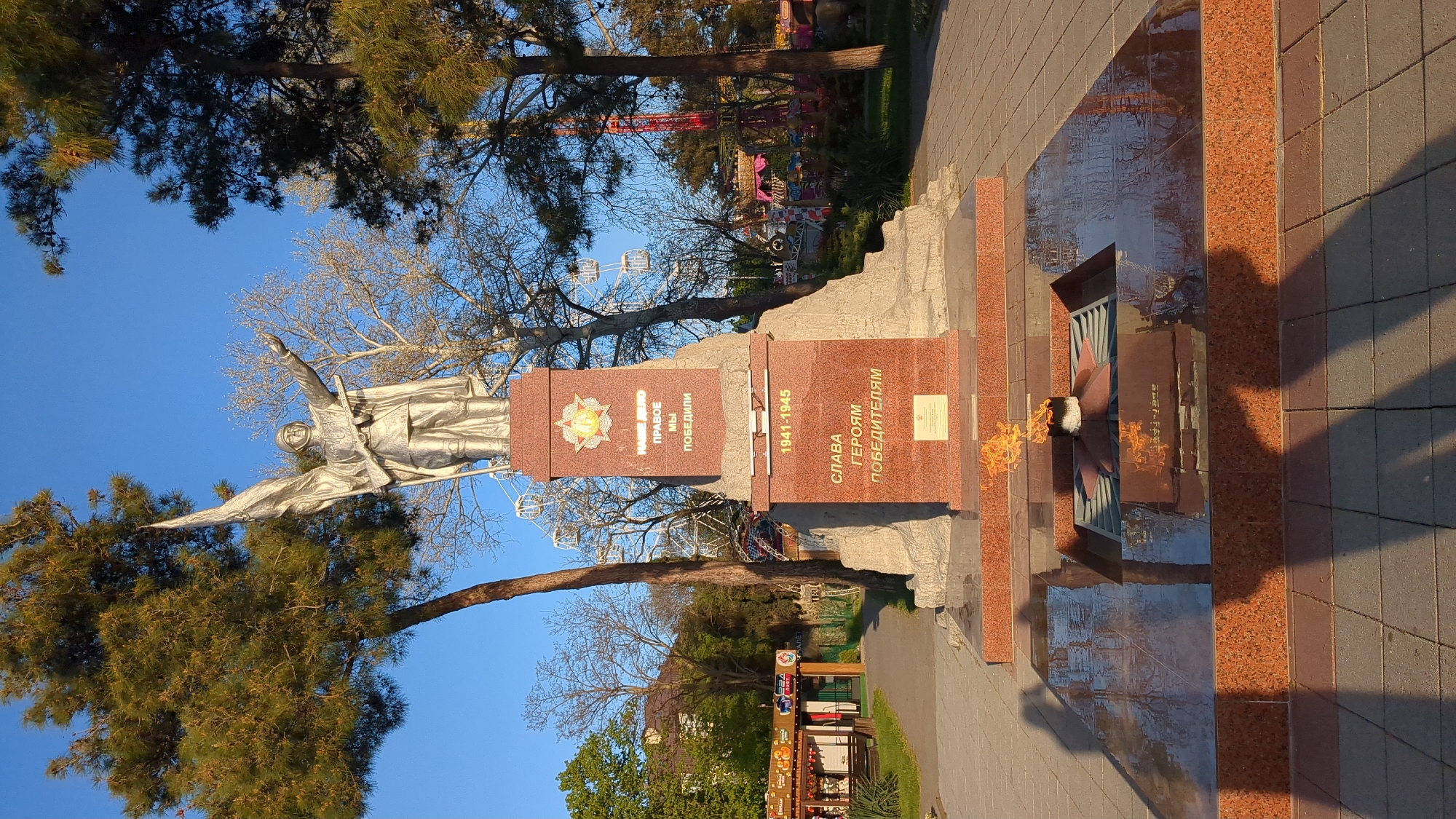 This media file captures an outdoor war memorial, likely situated in Gelendzhik, Russia. The central feature is a towering, silver-colored statue of a human figure, possibly an angel or a victorious soldier, with arms outstretched, positioned atop a polished red granite pedestal.
The pedestal features inscriptions. On one side, a star-like emblem is visible above the Cyrillic text "НАШЕ ДЕЛО ПРАВОЕ МЫ ПОБЕДИЛИ," which translates to "Our cause is just, we won." On the main face of the pedestal, the years "1941-1945" are clearly marked, referring to the Great Patriotic War, followed by "СЛАВА ГЕРОЯМ ПОБЕДИТЕЛЯМ," meaning "Glory to the heroes-victors."
At the base of the monument, an eternal flame burns within a star-shaped metal structure, set into a polished granite and tiled platform. This flame is a common feature of war memorials, symbolizing perpetual remembrance.
The setting is a park-like environment with several trees, some evergreen with dark foliage, and others deciduous with bare branches, suggesting early spring or late autumn. In the background, under a clear blue sky, an amusement park is visible, featuring a Ferris wheel and other colorful rides, creating a visual contrast with the solemn memorial in the foreground. Paved paths of grey and reddish tiles surround the monument. The bright sunlight casts distinct shadows, indicating either early morning or late afternoon.