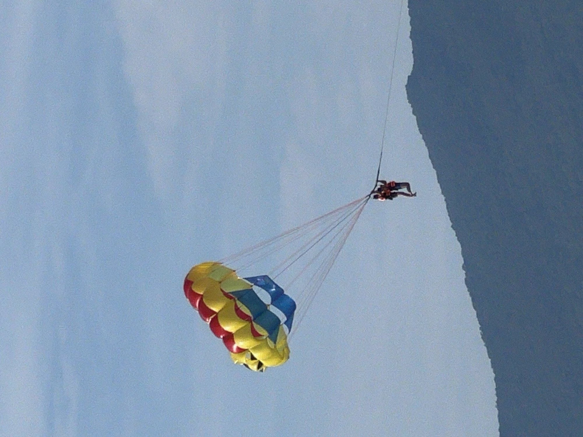 The image shows two people parasailing over a body of water with a mountainous coastline in the background. The parasail is large and colorful, with red, yellow, and blue sections. The two individuals are wearing orange life vests and are suspended beneath the parasail by ropes. The sky is a clear blue with some wispy clouds. The mountainous coastline is dark blue and covered in lush green vegetation. This scene is likely taking place near the beaches of Da Nang, Vietnam, known for their parasailing and other water sports.