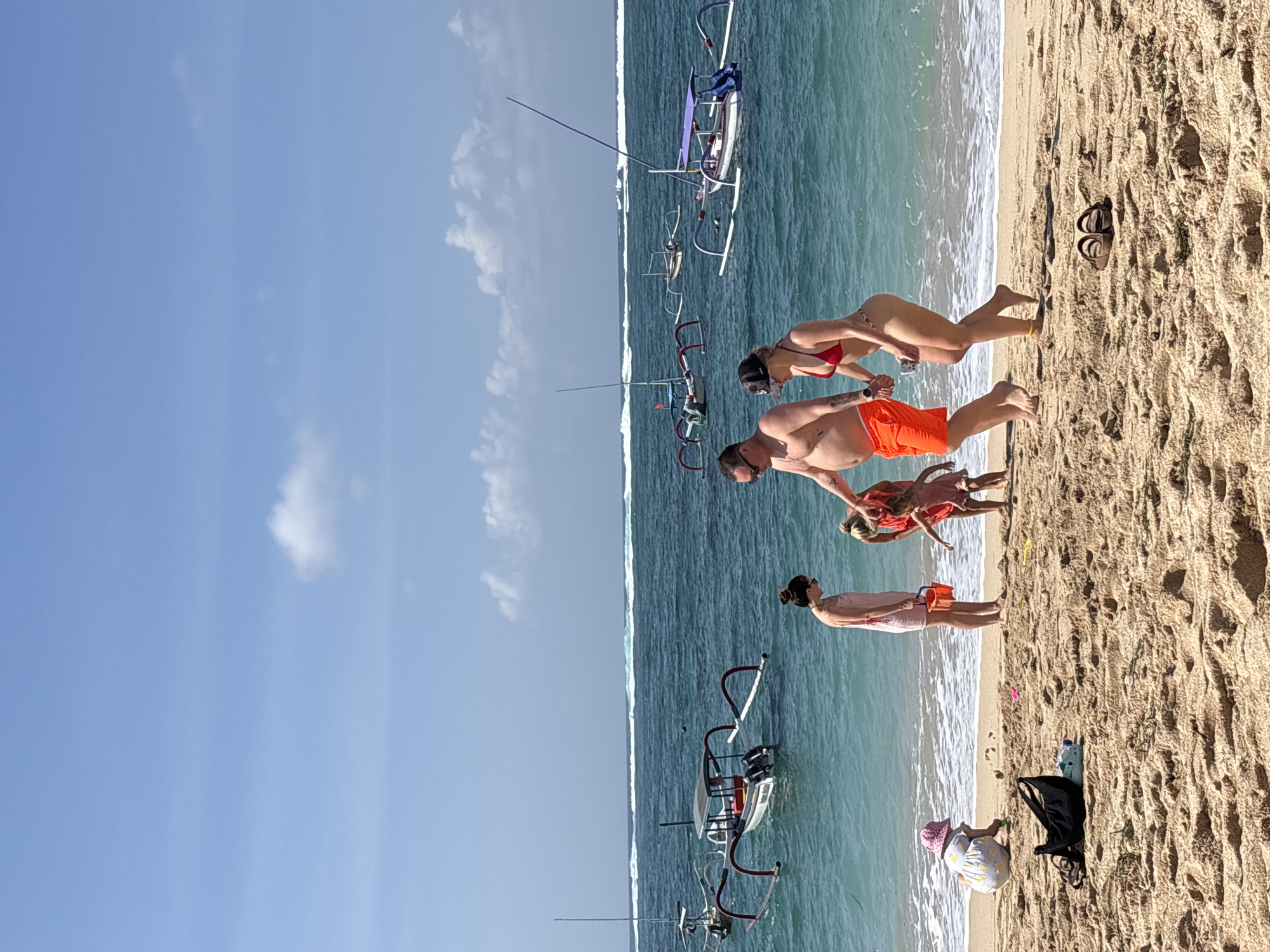 The image depicts a sunny day at the beach in Kuta Selatan, Indonesia. The foreground is dominated by sand, with a black bag and a pair of sandals visible. In the mid-ground, several people are present on the sandy shore. A man in orange shorts and a woman in a red bikini are walking hand-in-hand. Nearby, a woman in a pink dress and two young children are interacting. One child, wearing a yellow dress and a pink hat, is playing in the sand. The ocean stretches out behind them, with several traditional outrigger boats (jukung) floating on the turquoise water. Waves are breaking in the distance, and the sky is clear blue with a few white clouds.