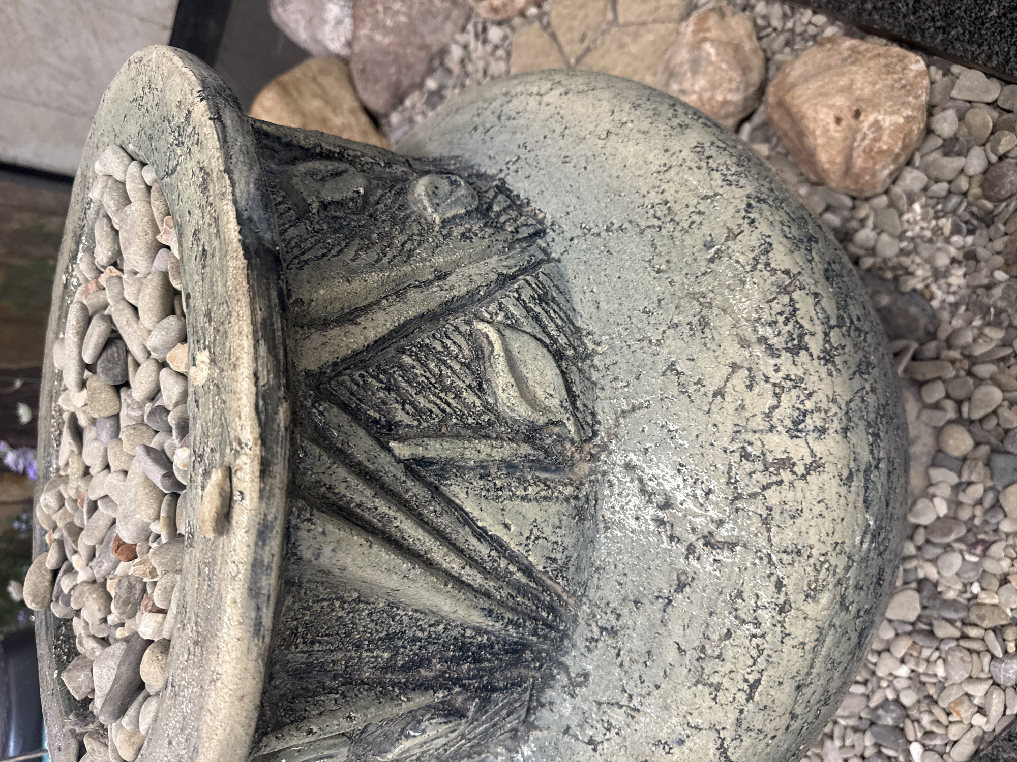 The image shows a close-up of a decorative ceramic pot filled with small pebbles. The pot has a textured, weathered finish with intricate carved designs on its body. It is surrounded by larger rocks and gravel. The scene appears to be outdoors, possibly in a garden or public space. There are no people or animals visible in the image. The location is specified as Yalta, Ukraine.