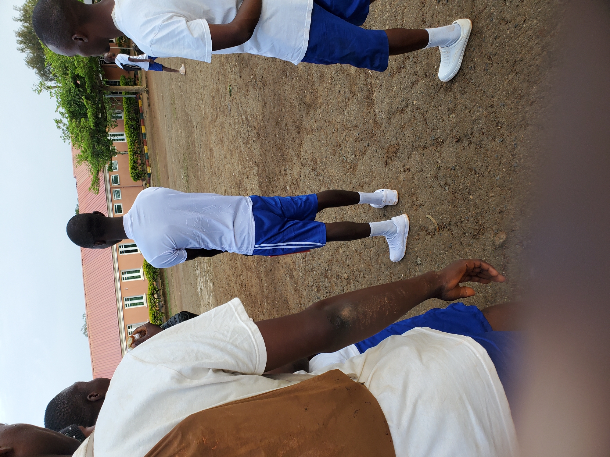 A group of young men, likely students, are gathered outdoors on a dirt ground. The central figure is a young man wearing a white t-shirt and blue shorts, with white socks and white sneakers. He is facing away from the camera and appears to be engaged in some activity. To his left, another young man, also facing away, wears a similar white t-shirt and blue shorts. To the right of the central figure, a third young man, dressed in a white t-shirt and blue shorts, is partially visible. In the foreground, a young man wearing a white t-shirt and blue shorts is seen from the side. The background features a building with a reddish-orange roof and several windows, suggesting an institutional setting. Trees and vegetation are also present, indicating an outdoor environment within or near a populated area. The overall scene depicts a casual gathering of individuals in what appears to be a school or dormitory compound in Kura, Nigeria.