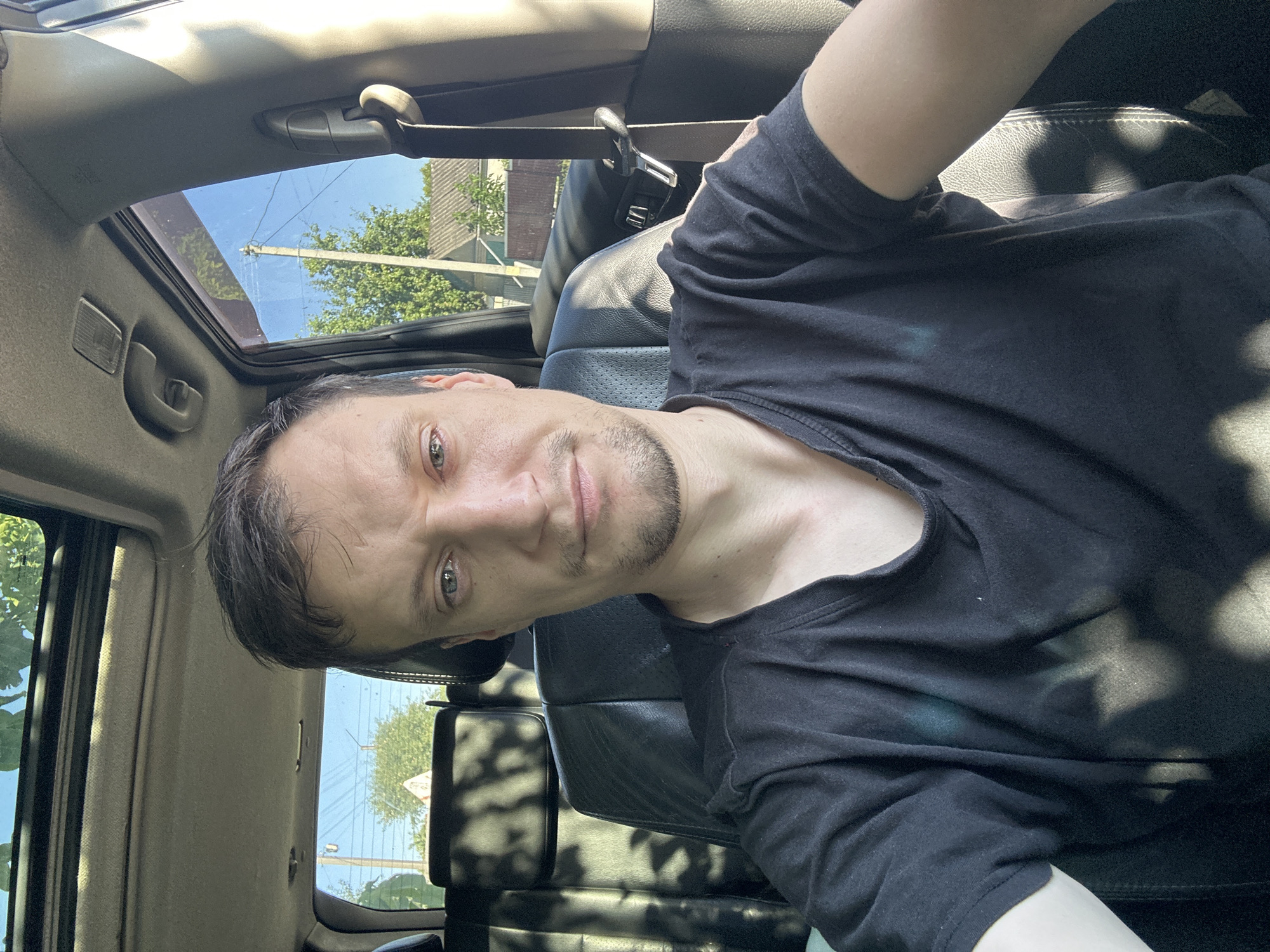 A man is seated inside a vehicle, taking a selfie. He has short, dark hair and is wearing a black t-shirt. The interior of the car is visible, including black leather seats and a sunroof. Sunlight streams through the sunroof and windows, casting shadows on the interior and the man's clothing. Outside the car, a residential area with houses and trees is visible under a clear blue sky. The location is identified as Shatava, Ukraine.