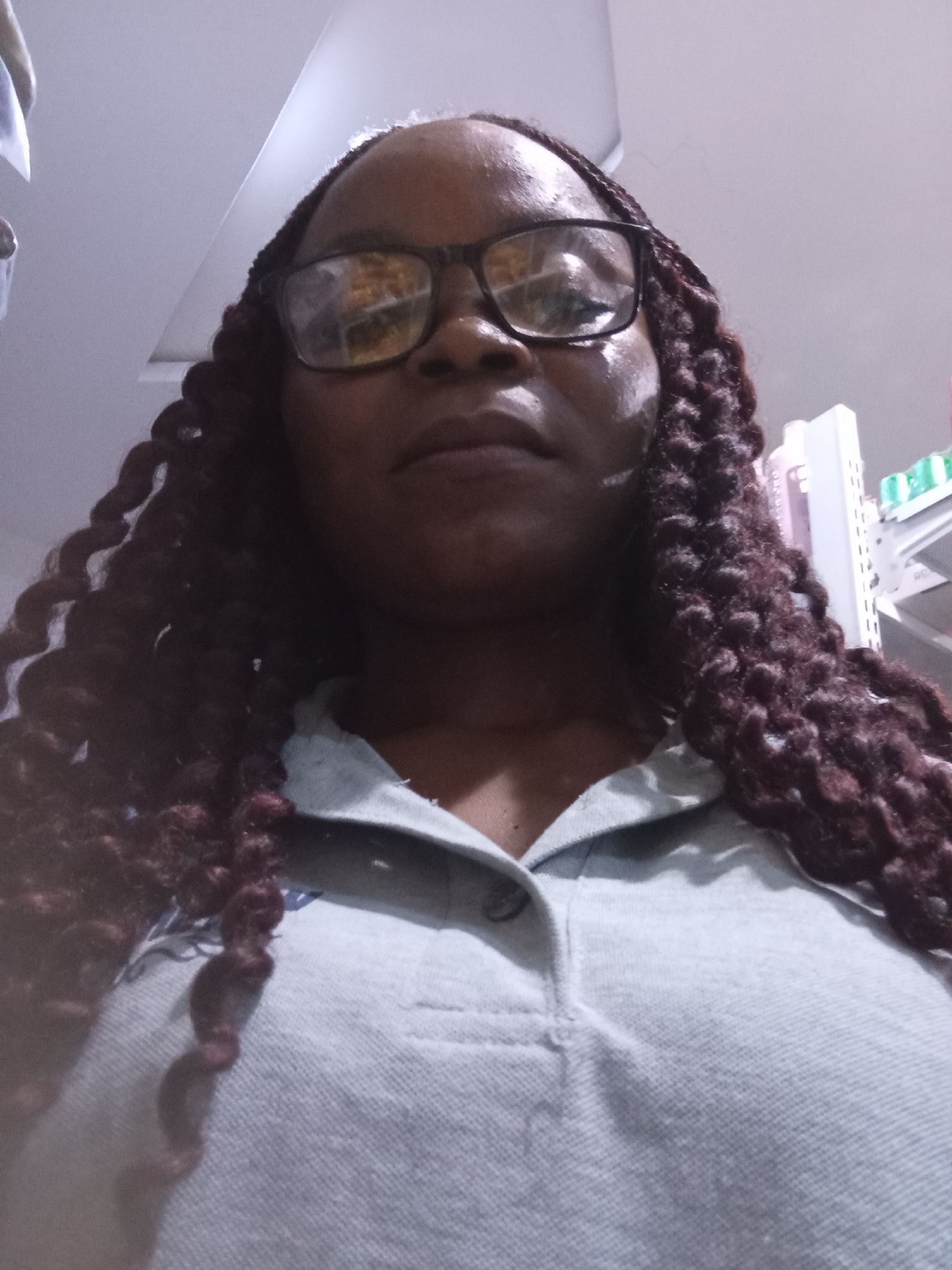 The image shows a close-up, low-angle shot of a Black woman wearing glasses and a light blue collared shirt. Her dark, braided hair is styled in thick twists that frame her face and extend down past her shoulders. The woman is looking slightly upwards, and her expression is neutral. The lighting suggests an indoor setting, possibly a well-lit room or a store, given the presence of what appear to be shelves with items in the background, though they are out of focus. The overall tone of the image is casual, likely a selfie. There is no visible text in the image. The provided location context is Howayi, Nigeria.