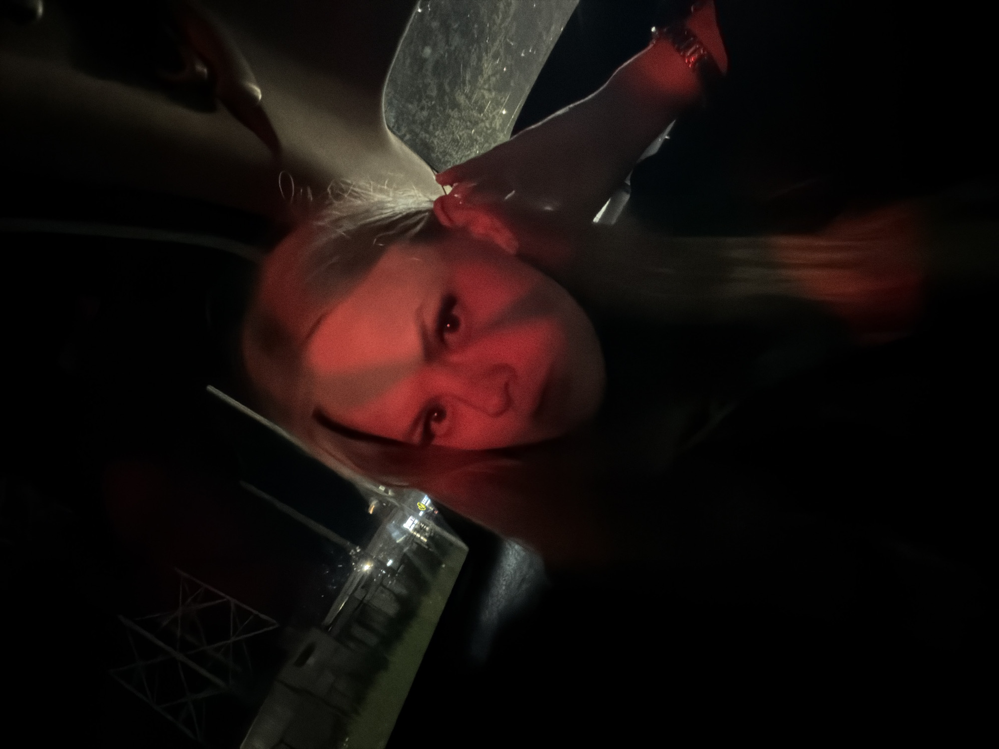 The image shows a young woman in a car at night. She has blonde hair and is illuminated by red light, casting a shadow across her face. Her right hand is near her ear, and she appears to be wearing a bracelet. Outside the car window, there are lights and structures that suggest an urban or industrial setting. The background is dark, with some blurry lights and what appears to be a power pylon. The overall impression is of a candid, perhaps selfie-style shot taken in a moving vehicle or at a standstill.
