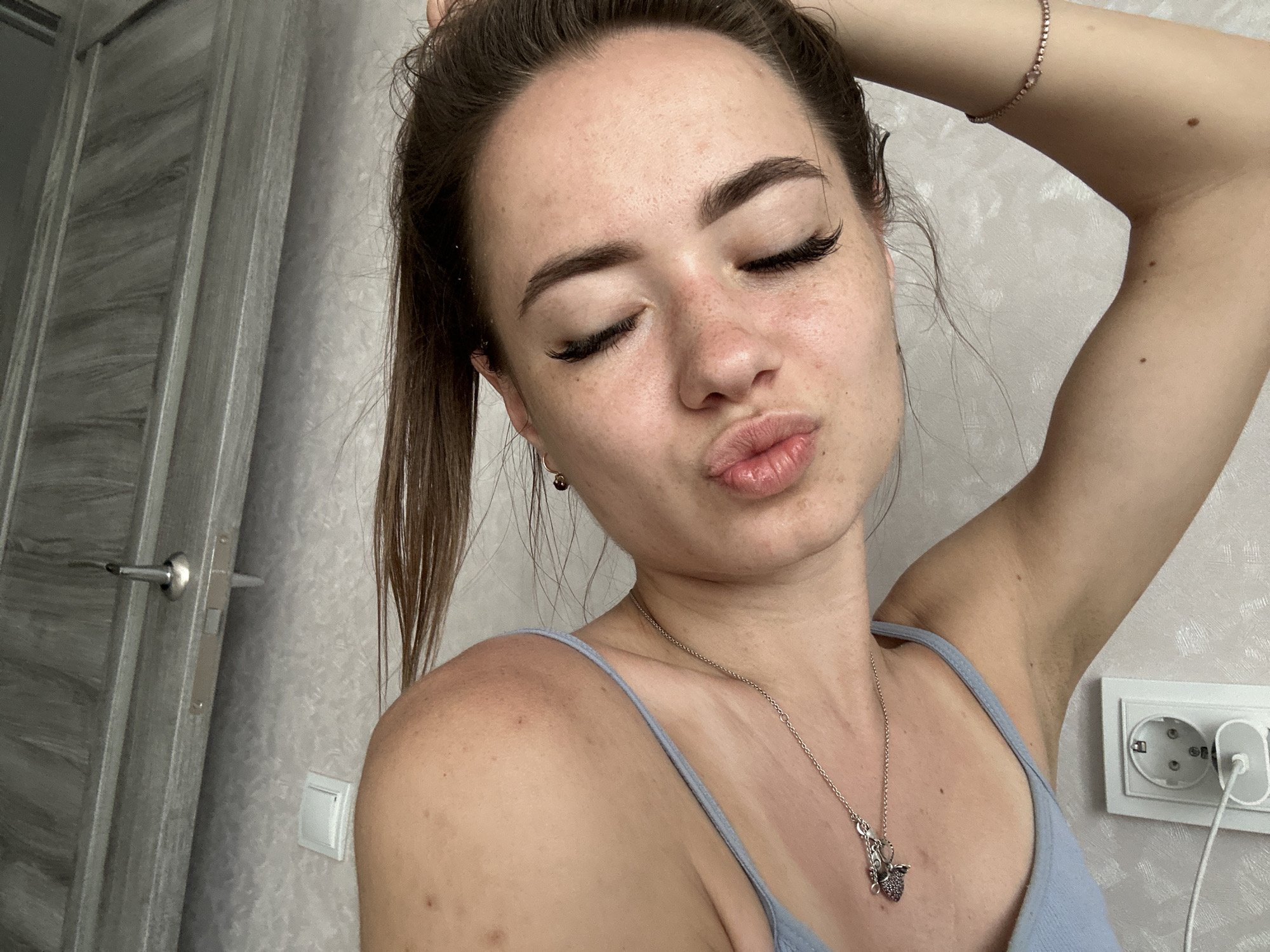 A young woman with dark brown hair pulled back in a ponytail is taking a selfie in what appears to be a bedroom. She is wearing a light blue tank top and a silver necklace with a pendant. Her eyes are closed, and her lips are pursed in a kissy face. Her right arm is raised and bent behind her head, revealing a diamond bracelet on her wrist. The background shows a gray door with a silver handle and a wall with textured wallpaper. There is also a white electrical outlet on the wall. The lighting is bright, and the image is in focus.