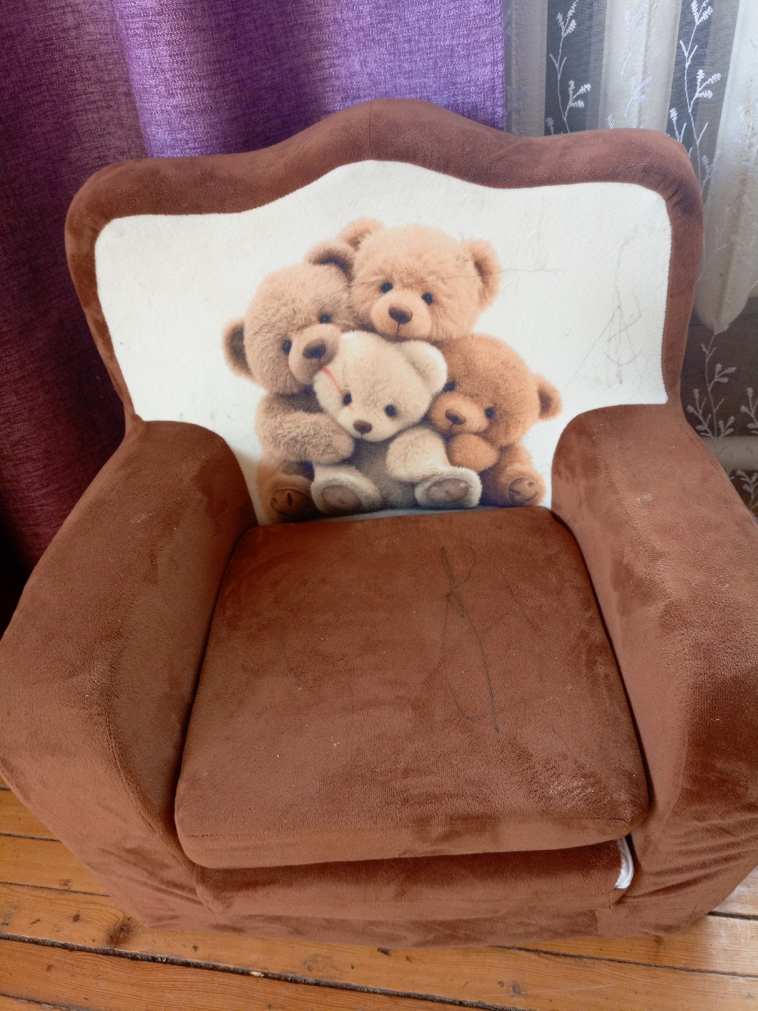 A brown, plush children's armchair with a printed image of three teddy bears is positioned on a wooden floor. The armchair has a high, curved backrest with a white panel featuring the teddy bear illustration. Behind the armchair, a purple curtain is visible on the left, and sheer white curtains with a floral pattern are on the right. The armchair appears to be in a domestic setting, possibly a living room or playroom. The image does not provide any information related to the city of Churapcha, Russia.