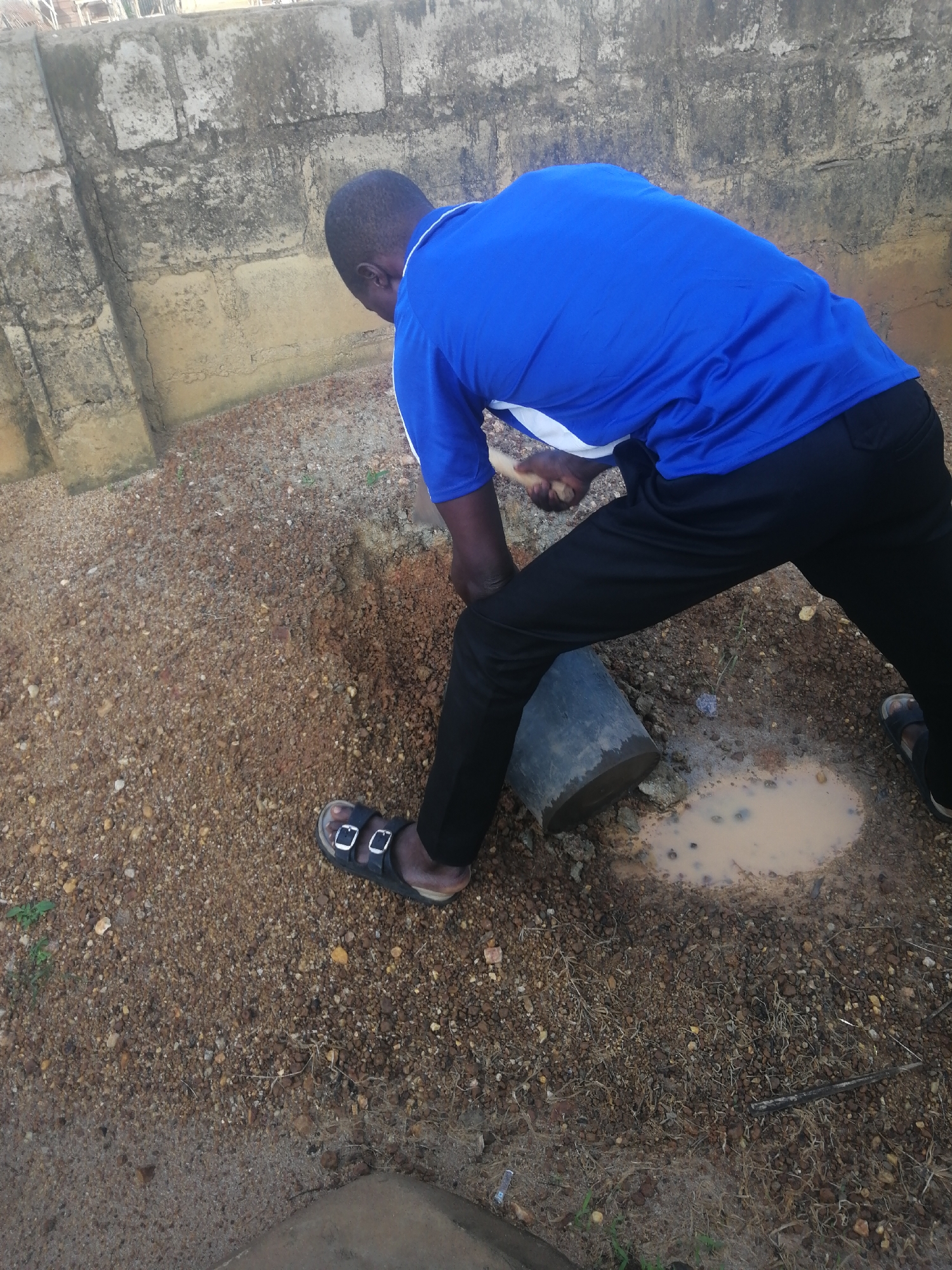 In Ajasse Ipo, Nigeria, a person is seen digging in the ground. They are wearing a blue shirt and black pants, with black sandals on their feet. A gray bucket is positioned to their right. The ground is covered in dirt and small rocks. A muddy puddle is visible near the bucket. A rough, textured wall made of concrete blocks forms the background. The overall scene appears to be an outdoor area, likely a yard or an open plot of land within the city.