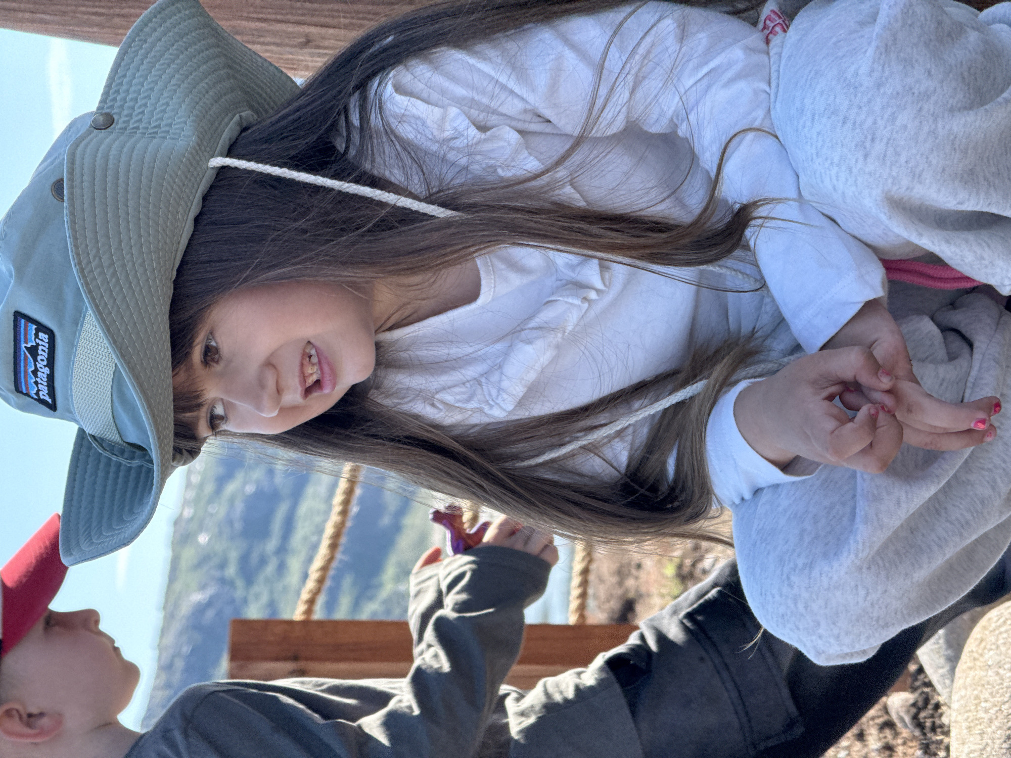 A close-up shot shows two children outdoors. On the right, a girl with long brown hair wears a light green bucket hat with a Patagonia patch and a white ruffled shirt. Her hands are clasped in her lap, and she looks to her left. On the left, a boy with short brown hair wears a red baseball cap and a dark gray hooded jacket. He is handing a small purple toy to someone out of frame. They are sitting on a rocky surface with a wooden railing and rope in the background. The landscape suggests an outdoor recreational area. The image does not contain information that would place it in Teriberka, Russia.