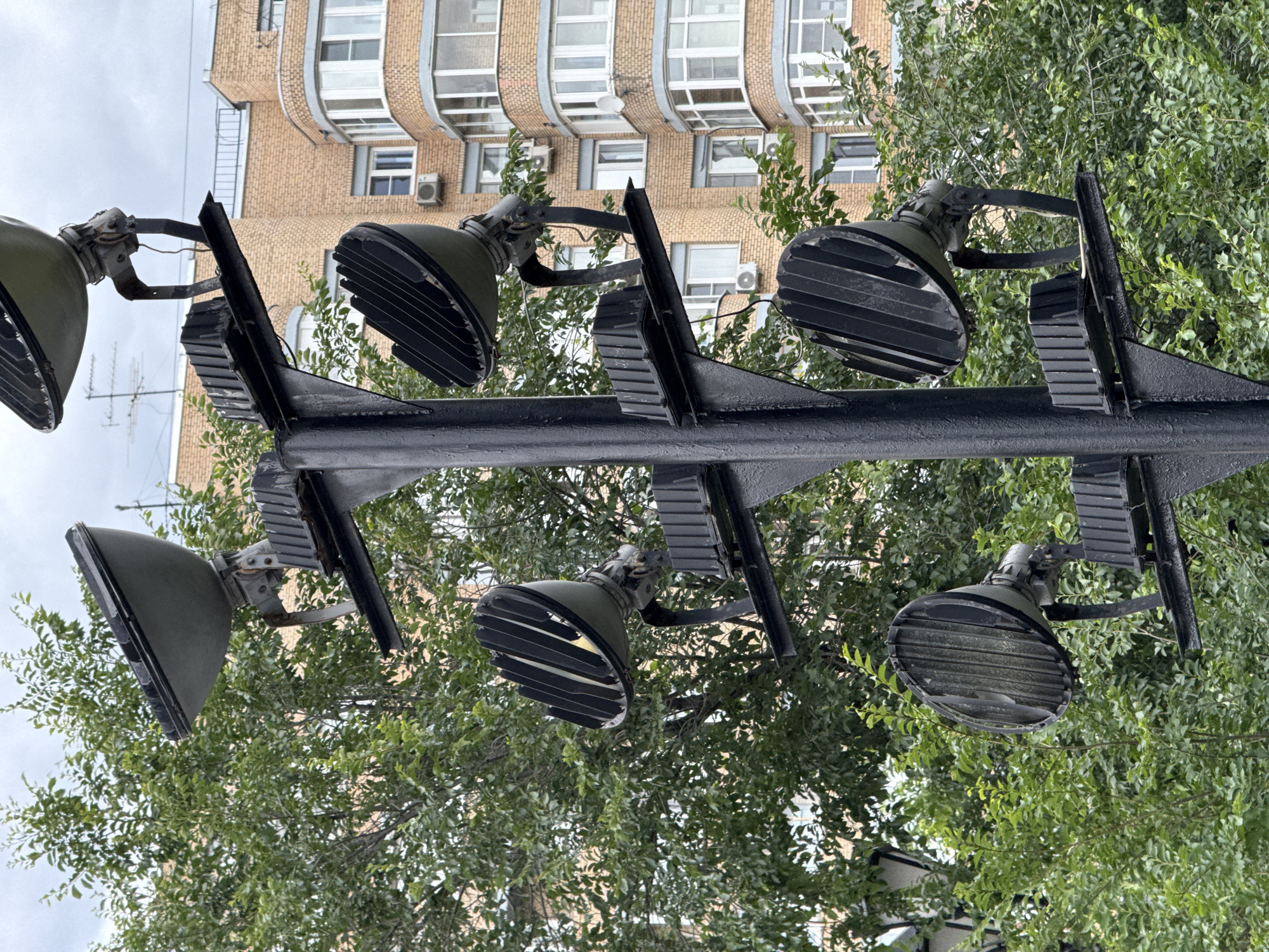 The image shows a vertical arrangement of six large, industrial-style spotlights mounted on a black metal pole. The spotlights are angled slightly downwards and to the left. They have a rugged, utilitarian design, with a green metal casing and a ribbed lens. Some of the lenses appear to be dusty or weathered.
In the background, a multi-story brick building with many windows is visible. The building has a curved facade and appears to be modern or from the mid-20th century. Air conditioning units are mounted on the exterior of the building. Dense green foliage from trees surrounds the spotlights and partially obscures the building. The sky is overcast, suggesting a cloudy day.
The scene appears to be an outdoor urban setting, likely in a park, plaza, or along a street where these lights would be used for illumination. There are no people visible in the image, and no discernible activity is taking place. The lighting is natural daylight, and the weather appears to be mild and overcast. The image is shot from a low angle, looking up towards the spotlights and the building. No text is visible in the image. The overall impression is one of functional infrastructure in a natural and built environment.