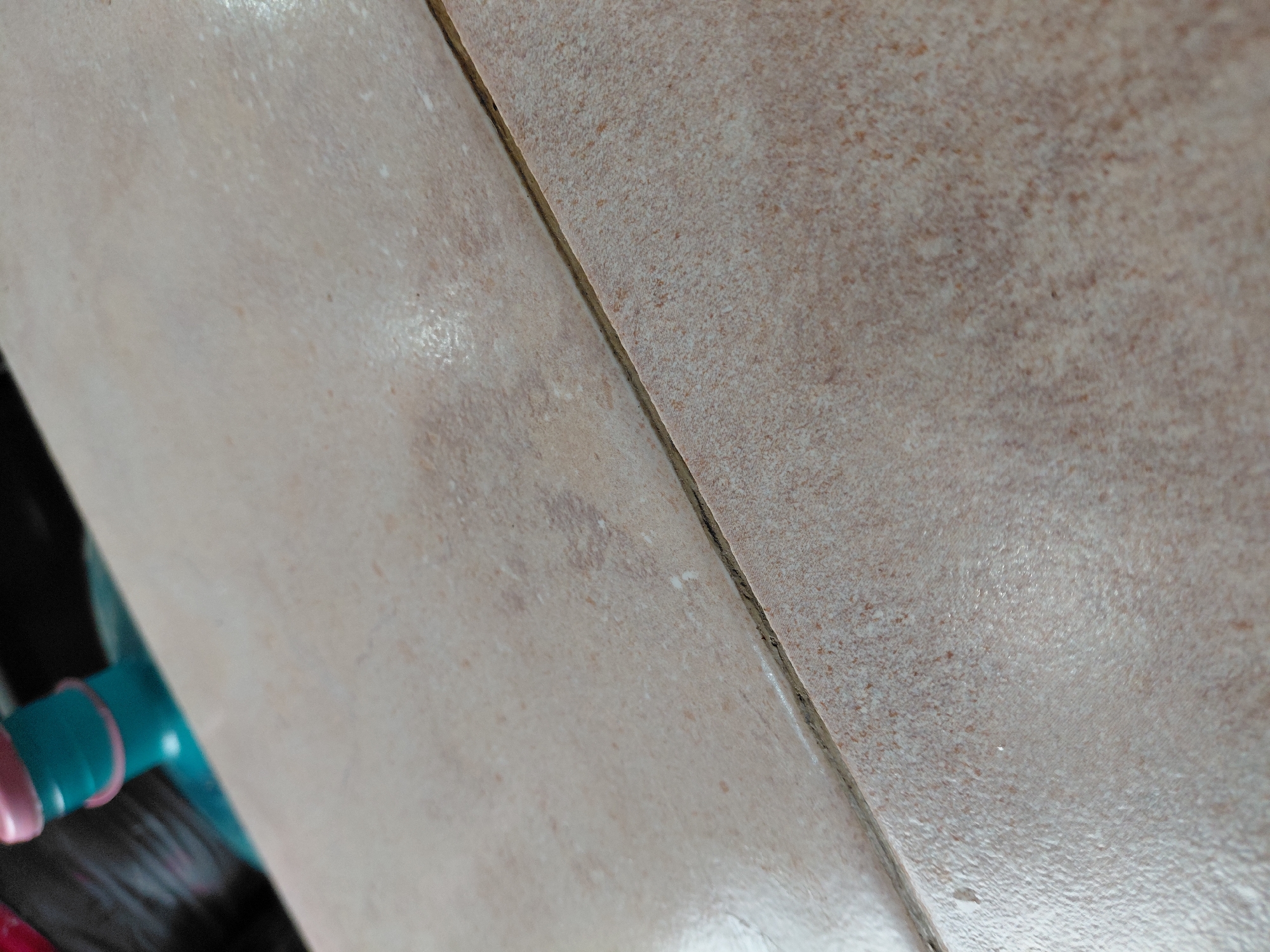 A close-up, downward-angled photograph captures two light-colored ceramic or porcelain floor tiles separated by a dark grout line. The tiles are predominantly a light beige or sandy brown hue, featuring a mottled, textured surface with subtle variations in color, including scattered darker specks and irregular lighter, almost white, patches. The right tile exhibits a noticeably coarser, more granular texture, while the left tile appears slightly smoother and shows some reflective sheen in parts.
The grout line, running diagonally from the top center to the bottom left, is dark gray or black, providing a strong contrast against the lighter tiles. It appears somewhat worn, with visible dirt or discoloration in sections along its length.
In the bottom-left corner of the image, partially visible and out of focus, is a bright teal-colored plastic object, around which a distinct pink band is wrapped. This object seems to be resting on or adjacent to the tiled surface.
The scene depicts an indoor setting, likely a floor, with no specific activity taking place. The overall impression is one of a domestic or commercial interior. No text is visible within the frame. The location context provided is Santa Ana, Venezuela.