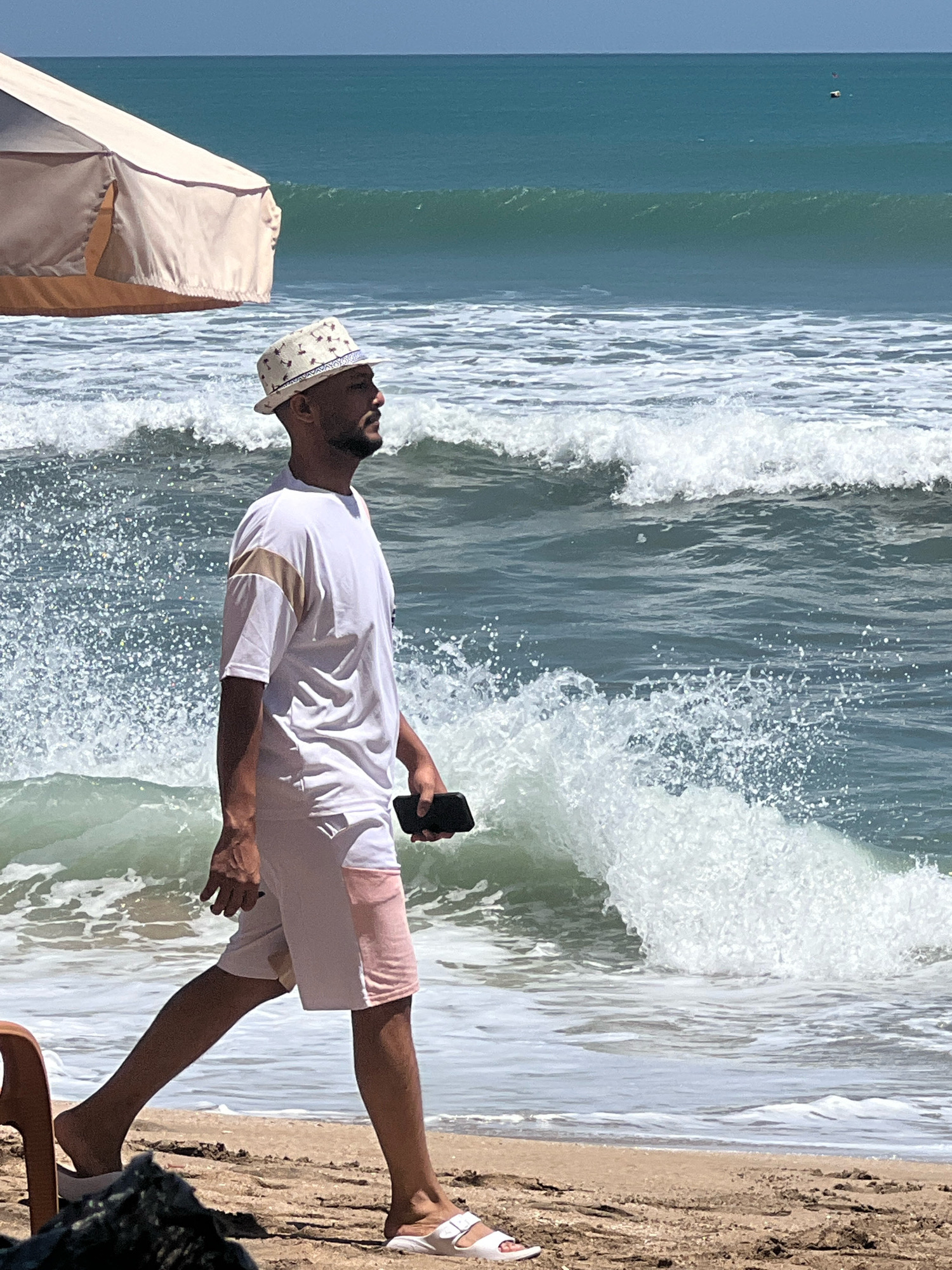 A man in a white t-shirt and patterned hat walks along a sandy beach in Kuta, Indonesia, with the ocean and waves in the background. He is holding a black smartphone in his right hand. The man is facing to the right, with his profile visible. He is wearing white shorts with a pink panel. A beach umbrella is partially visible on the left side of the image. The water is a clear blue-green, with white foam from the breaking waves. The sky is a bright blue.