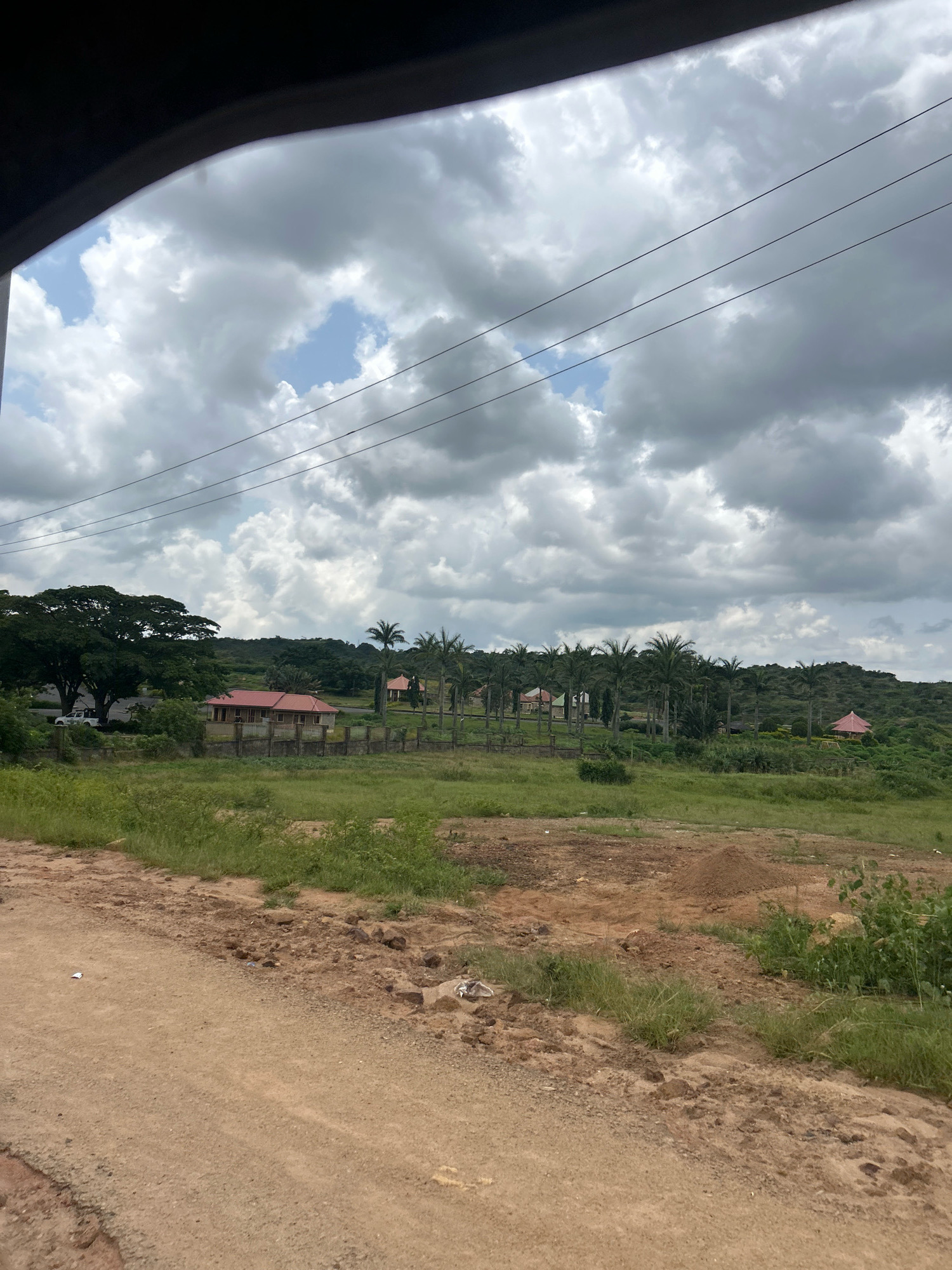The image depicts a residential area on the outskirts of Jos, Nigeria, characterized by a mix of vegetation and man-made structures under a cloudy sky. In the foreground, a dirt road leads into the scene, with sparse green grass and patches of exposed earth. Further into the image, a low concrete fence encloses a property with a red-roofed building. Beyond this, a line of palm trees stands tall, interspersed with other trees and vegetation. In the distance, rolling hills covered in greenery form the backdrop. The sky is filled with heavy, grey clouds, suggesting an overcast or impending weather event. A white vehicle is parked near the large tree on the left. Power lines are visible stretching across the upper portion of the frame. The overall impression is of a developing or semi-rural residential setting in Jos.