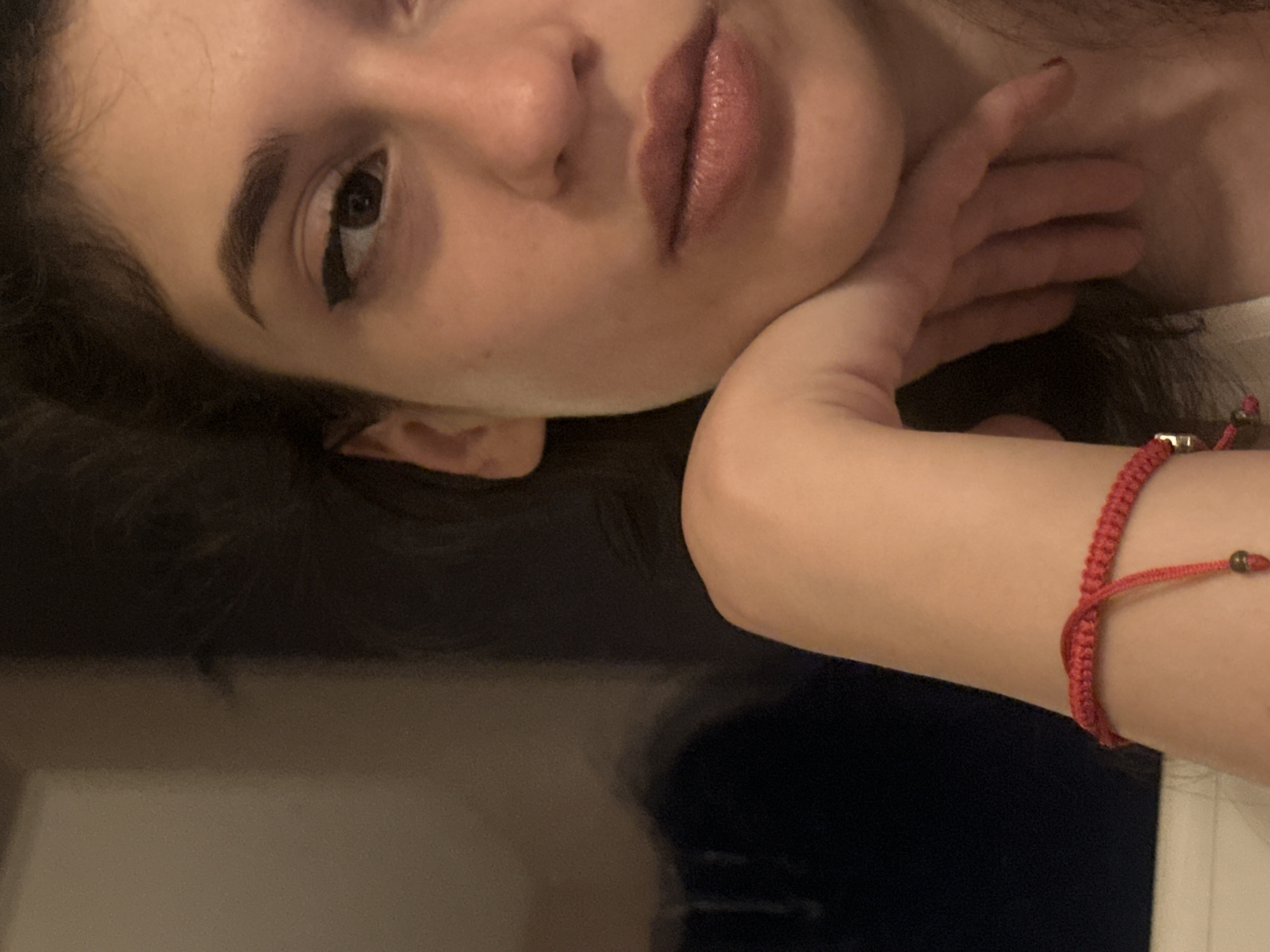 The image is a close-up, candid shot of a young woman. She has dark hair, dark eyes, and is wearing winged eyeliner. Her left hand is resting on her chin, and she is wearing a red braided bracelet on her left wrist. The background is blurred, with a dark blue area on the left and a light brown area on the right. The lighting suggests an indoor setting, possibly at night. There are no discernible landmarks or elements that would allow for the identification of the city of Sofia, Bulgaria, or any specific location within it.
