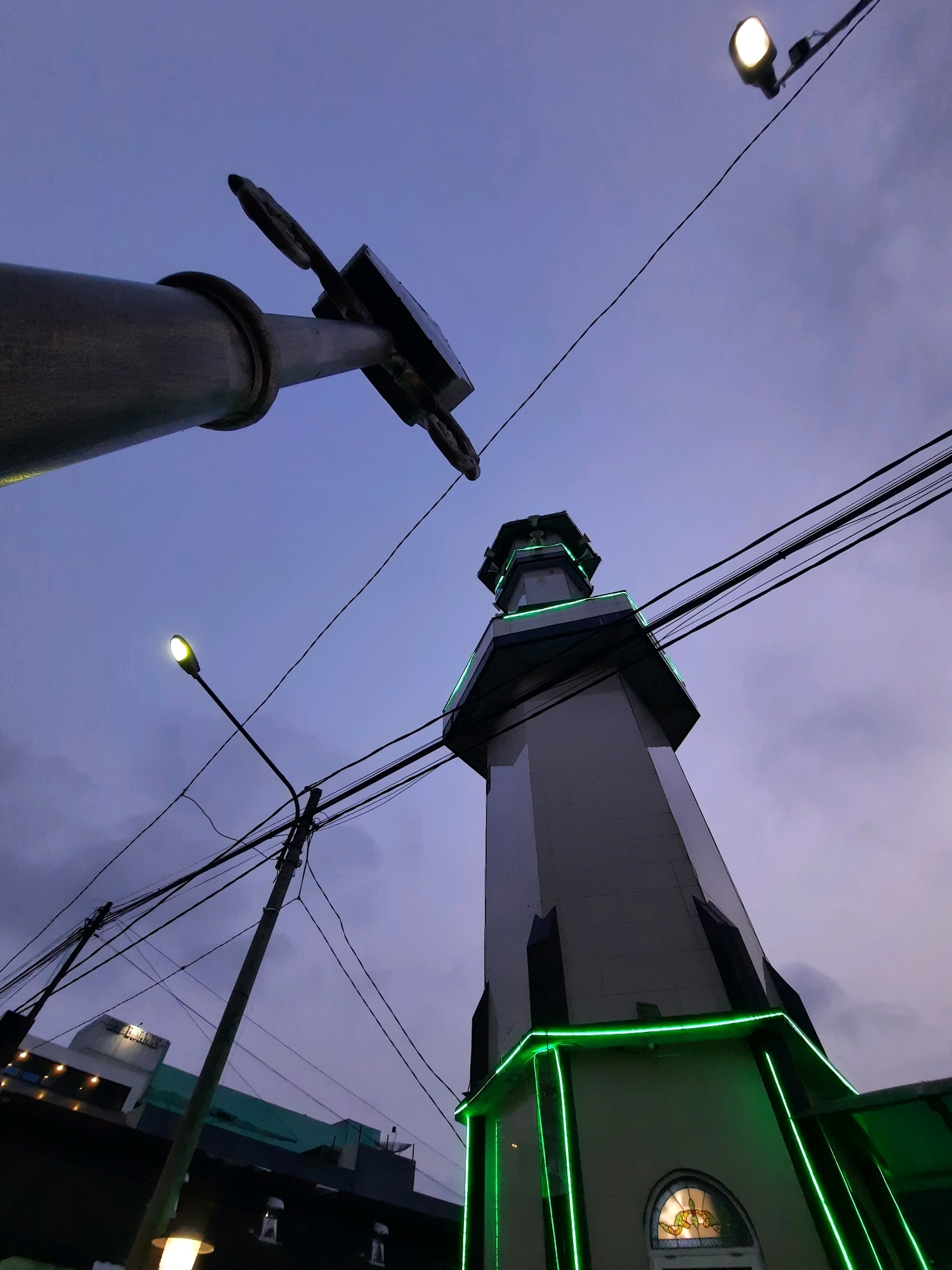 The image, taken at dusk in Kutoarjo, Indonesia, focuses on a mosque minaret and a utility pole against a darkening sky. The minaret, a tall, octagonal structure, is illuminated with green neon lights along its upper edges. It features small windows and a decorative stained-glass element above an entrance. Several black electrical wires crisscross the sky, obscuring parts of the minaret and a distant building. To the left, a large cylindrical object, possibly part of a street lamp or a larger structure, dominates the foreground, angled upwards. Another street lamp with a white light source is visible in the upper right quadrant. In the lower left background, parts of buildings with illuminated windows and signage are faintly visible. The overall impression is an urban night scene with a blend of religious architecture and functional infrastructure.