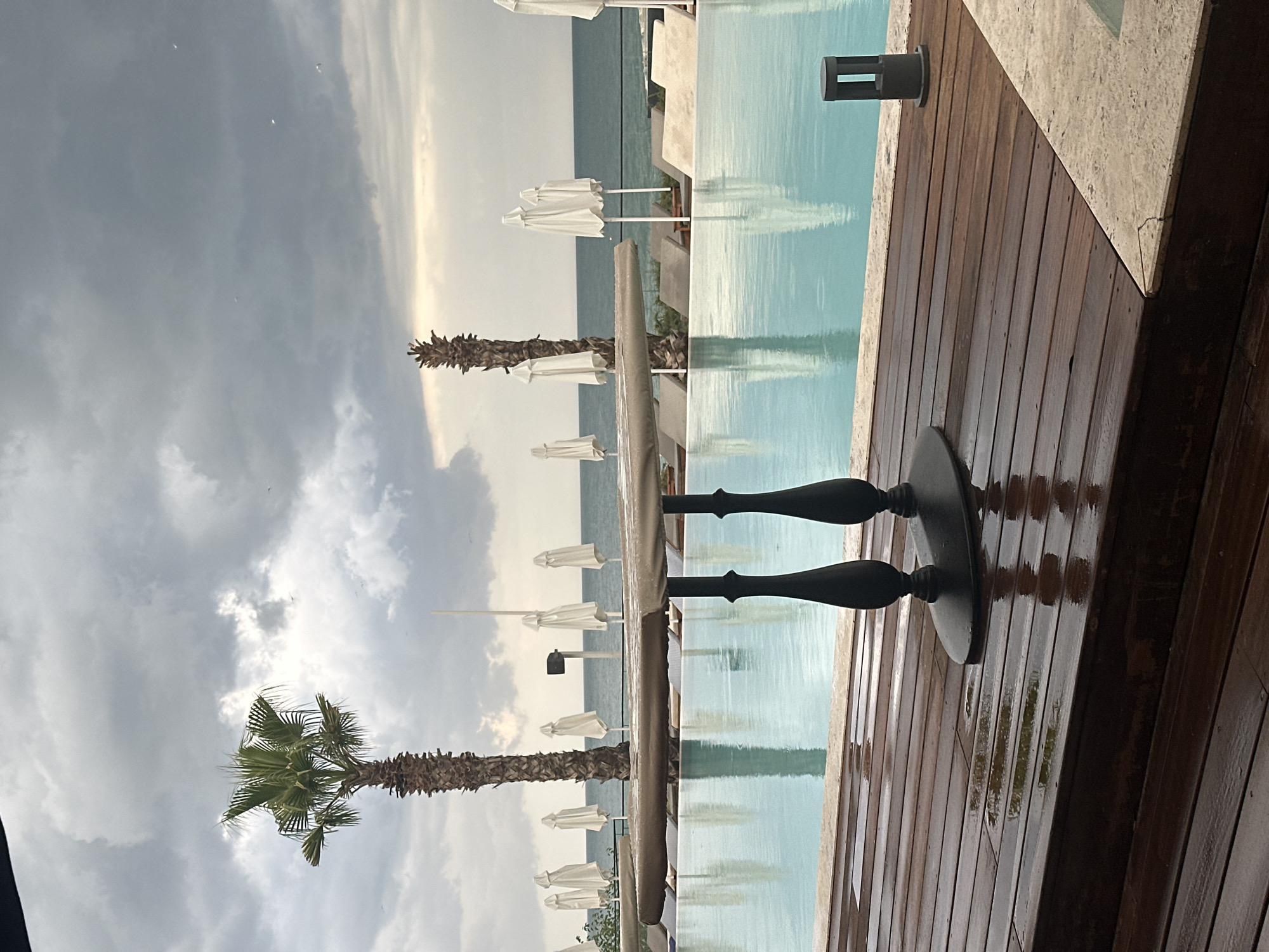 The image shows a partially covered table and chairs in the foreground, positioned on a wet wooden deck next to a swimming pool. Beyond the pool, lounge chairs and closed white umbrellas are visible, with the sea and a cloudy sky in the background. There are no people present in the scene. The setting appears to be an outdoor area at a resort or hotel, likely before or after a rain shower given the wet surfaces.