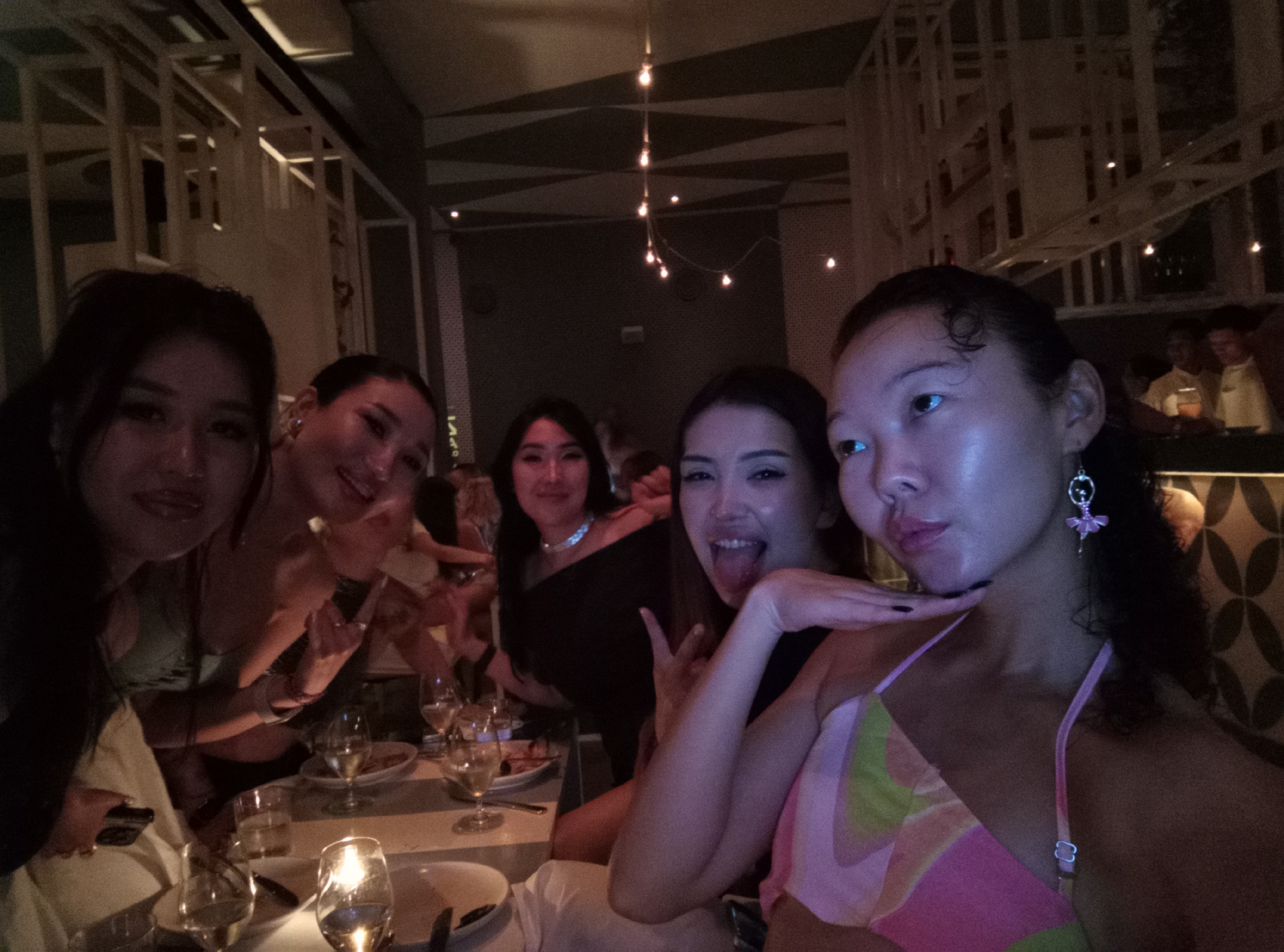 The image shows a group of five young women posing for a selfie in a dimly lit indoor setting. They are seated at a table with plates, glasses, and cutlery, suggesting a restaurant or bar setting. The person taking the selfie is on the right and wears a pink and green patterned top with ballerina earrings. The woman to her left is sticking her tongue out and making a peace sign. Another woman in the center of the group is wearing a black top and a necklace. The background includes geometric patterns on the ceiling and decorative wall panels, along with dim hanging lights. Based on the metadata, the location is North Kuta, Indonesia.