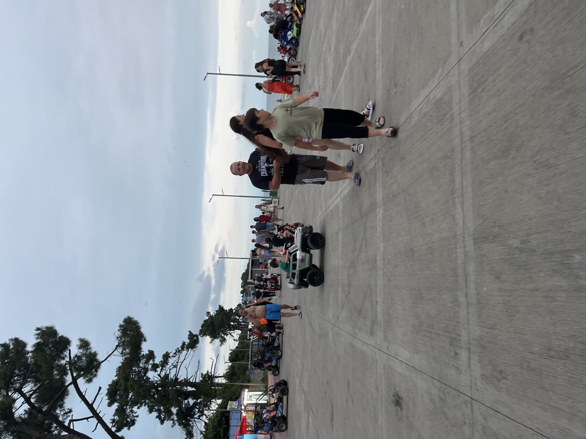 The image depicts a paved promenade in Ureki, Georgia, with a cloudy sky overhead. Several people are present, including adults and children. In the foreground, a young boy is seated in a toy car. To his left, a man in blue shorts and a black t-shirt is walking and talking on his phone. Behind them, a family is walking, with a woman with long dark hair and a man in a light-colored t-shirt and black pants. Further back, there are several parked go-karts and a variety of motorcycles. In the distance, more people are walking along the promenade. Trees are visible on the left side of the image, and the sea is hinted at in the background. The overall atmosphere appears to be a recreational area, possibly a beachfront promenade, with various forms of entertainment available.