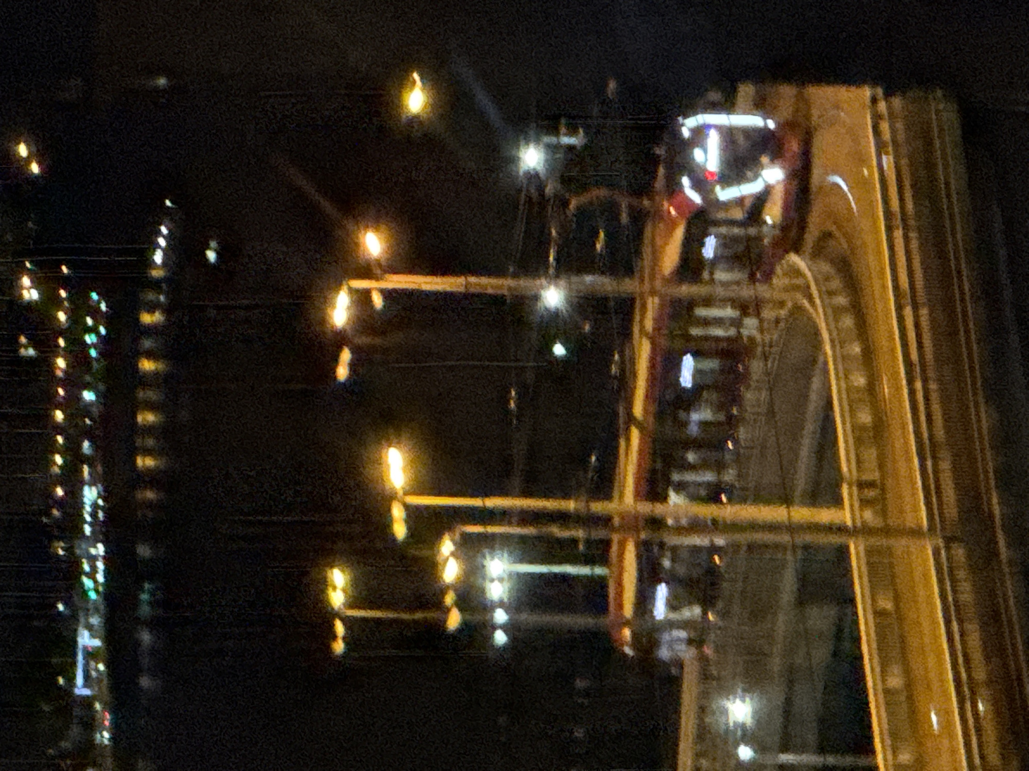 An urban scene at night, with a tram on a curved track illuminated by streetlights. The tram is red with white lights on its front. The track is bordered by a raised walkway with railings, all illuminated by yellow streetlights. In the background, across a body of water reflecting city lights, a row of buildings with illuminated windows is visible. The overall image is grainy due to low light conditions.