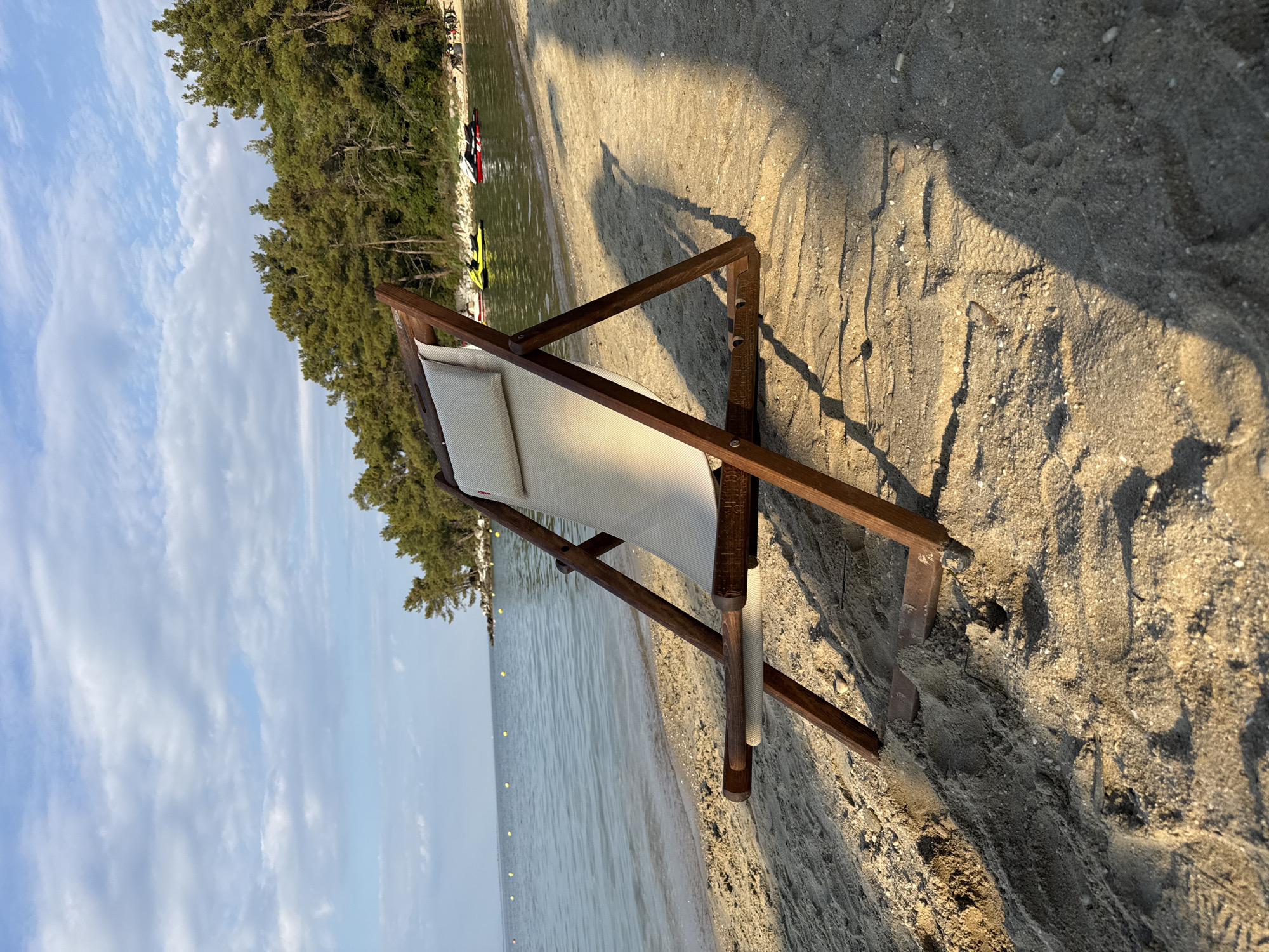 A wooden beach chair sits on a sandy beach in Thassos, Greece, facing the calm sea. The chair is made of dark wood and has a light-colored fabric seat and a small cushion. The sand is a light brown color, with some small pebbles and shells visible. In the background, the sea is a clear blue, with a few yellow buoys marking a boundary. A line of green pine trees borders the beach, and beyond them, the sky is a pale blue with scattered white clouds. Several jet skis are visible on the water in the distance, and a few people are present near the water's edge. The sun is casting long shadows across the sand, indicating it is likely late afternoon or early morning.