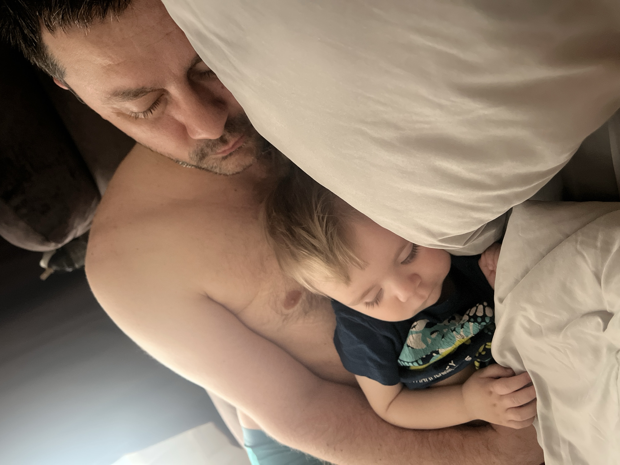 A man and a toddler are sleeping in bed. The man is fair-skinned and shirtless, with dark hair and a stubbled beard. He is lying on his side, facing the camera, with his eyes closed. The toddler is nestled in the man's arms. The toddler has blond hair and is wearing a dark blue t-shirt with a green dinosaur print. The toddler is also facing the camera, with their eyes closed. A white pillow is positioned above the man's head, and a white blanket is partially covering the toddler. The scene appears to be indoors, in a dimly lit bedroom. The location of Noviy, Russia, cannot be verified from the visual data.