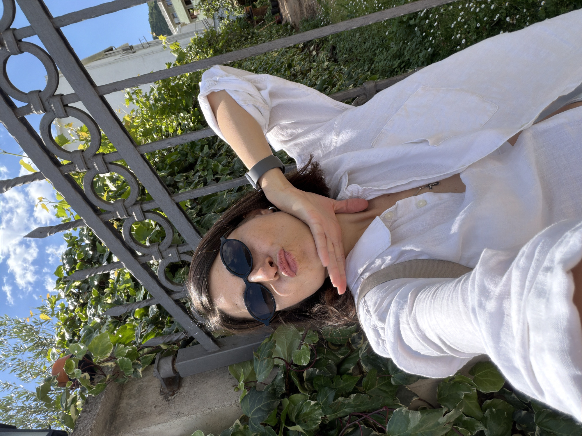 The image shows a woman lying on a metal park bench in Budva, Montenegro. She is wearing a white linen shirt and black sunglasses. She is holding her hand up to her face. The bench is surrounded by ivy. In the background is a white building with greenery. The sky is blue with some clouds.