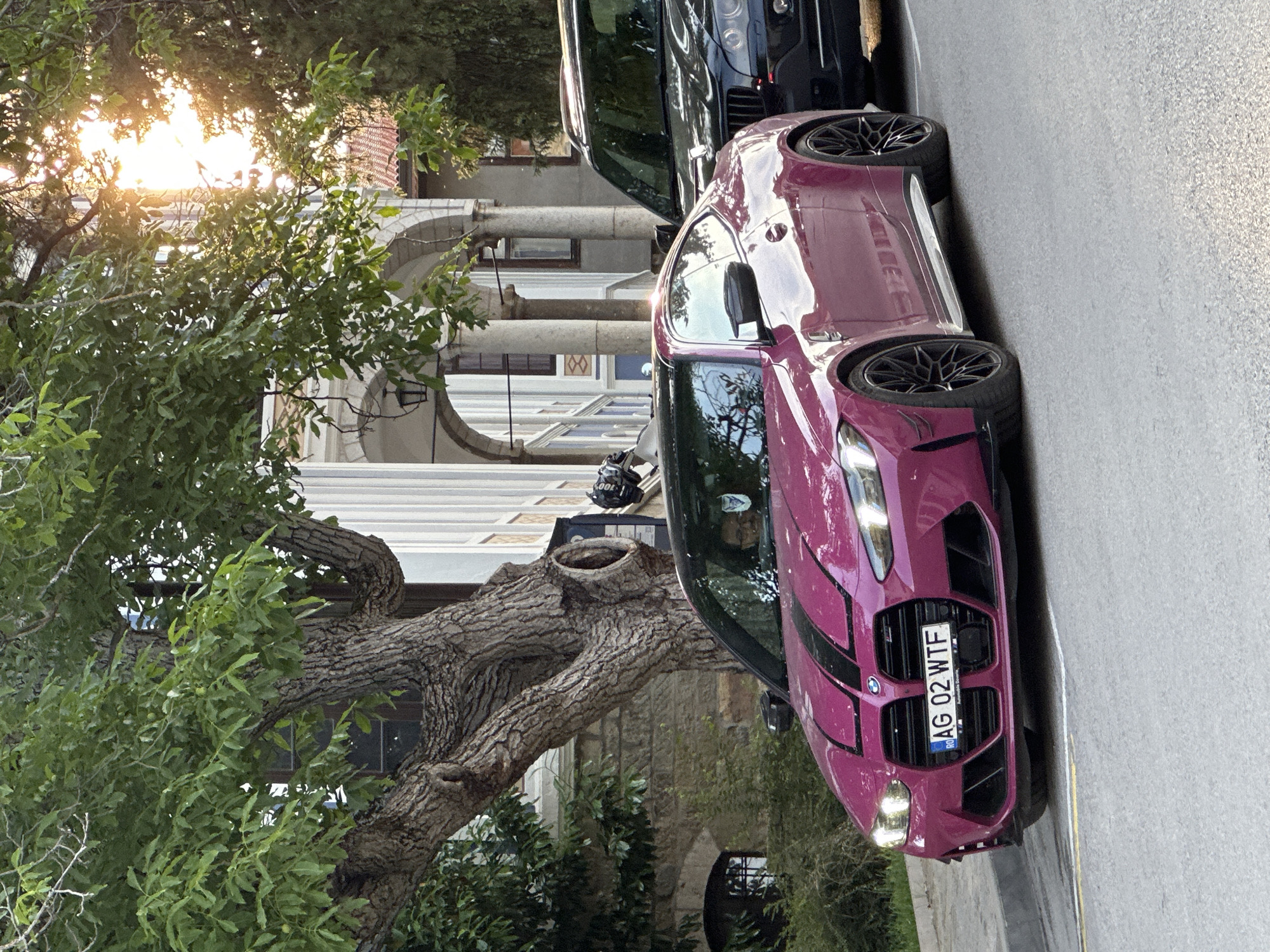 In the city of Veliko Tarnovo, Bulgaria, a striking pink BMW M4 coupe is parked on a street, with its front facing the viewer. The car has black racing stripes running down its hood. The license plate is visible, reading "RO AG 02 WTF". To the right of the BMW, a black SUV is also parked. Behind the vehicles, a building with classical architectural elements, featuring arches and columns, is visible. A large, mature tree with textured bark and lush green leaves stands prominently in the foreground, partially obscuring the building. Sunlight streams through the tree's foliage, creating a warm, dappled effect. The scene appears to be an outdoor street setting in a historic part of the city.