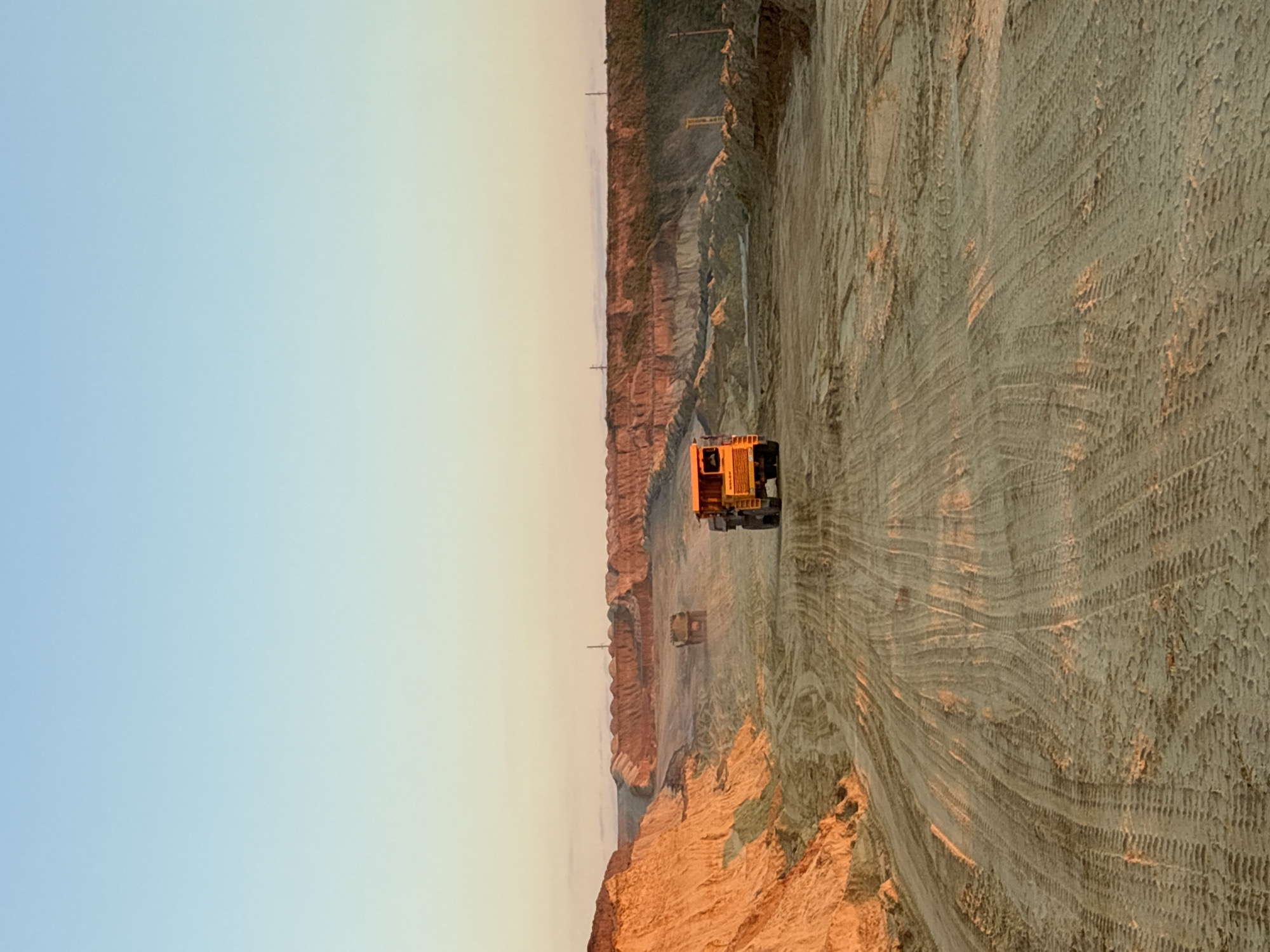 The image depicts an outdoor scene at a quarry or construction site, likely in Tatsinskaya, Russia, during what appears to be either sunrise or sunset, given the warm, golden light illuminating the sandy terrain and the clear, pale blue sky. The foreground is dominated by a wide, sandy track imprinted with tire tracks from heavy machinery. In the middle ground, two large, yellow dump trucks are visible. One is closer to the right side of the frame, and another, slightly smaller truck is further in the distance, positioned in the center of the track. The background features layered, sculpted mounds of earth and sand, characteristic of a quarry or excavation. Power lines and poles are intermittently visible along the upper edge of the distant mounds, suggesting proximity to infrastructure. The overall impression is one of an industrial or earthmoving operation in a vast, open environment, bathed in the soft glow of low-angled sunlight.