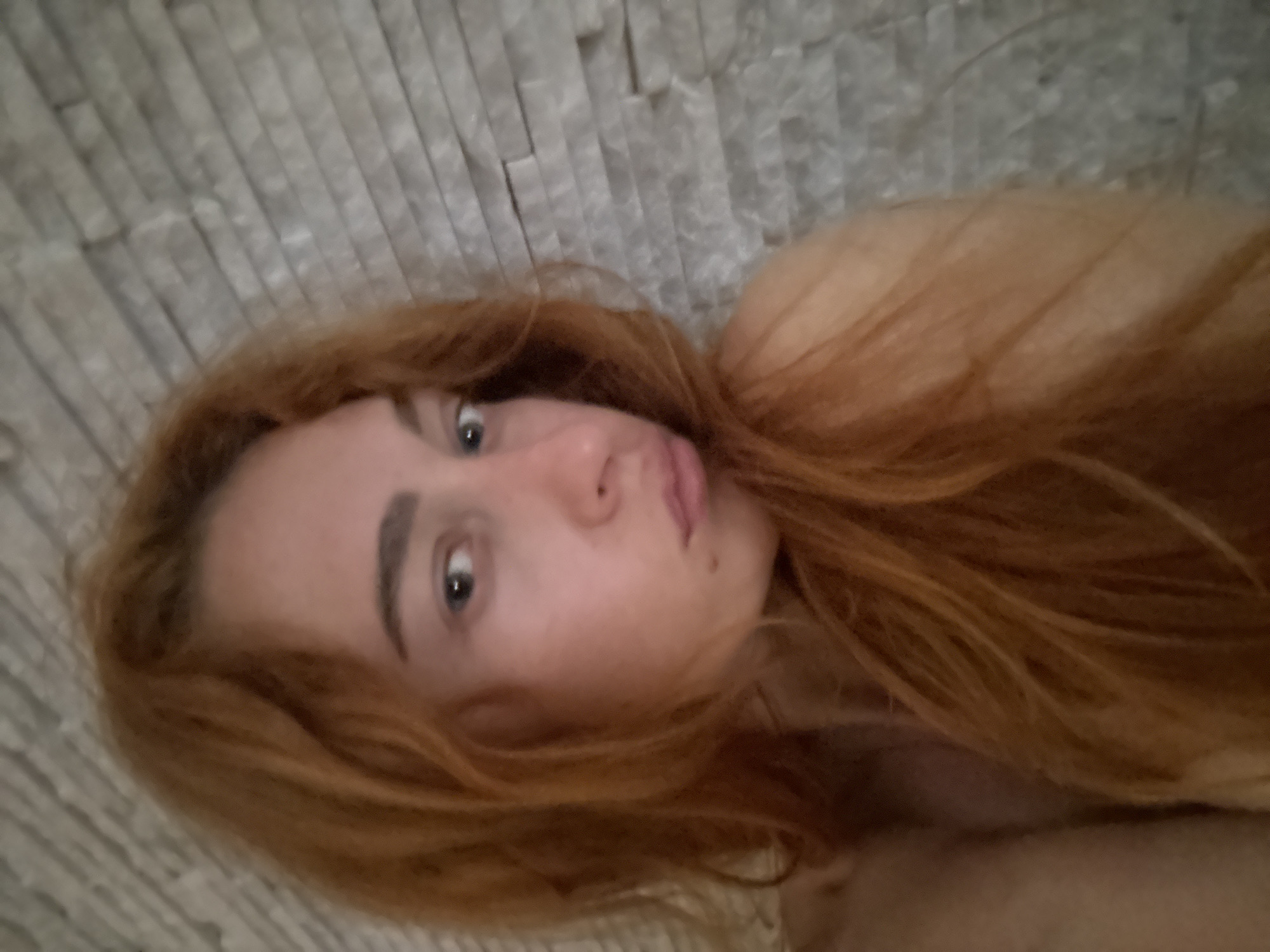 A woman with fair skin, red hair, and blue eyes is lying down on what appears to be a bed or soft surface. She is positioned with her head resting against a wall constructed of light-colored, rectangular stone bricks. The image is taken from a close-up, slightly angled perspective, focusing on her face and upper body. The setting does not provide enough information to determine a location in Budva, Montenegro.