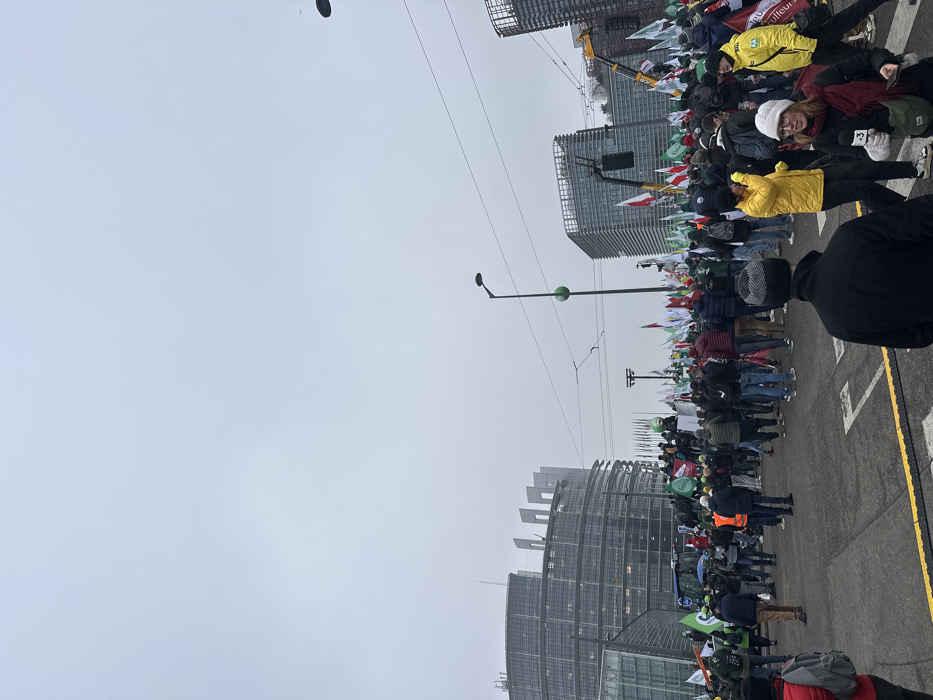 The image depicts a large outdoor gathering on a paved street in Strasbourg, France, positioned directly in front of the European Parliament's Louise Weiss building. The sky is uniformly overcast.
Numerous individuals, dressed in winter coats, hats, and scarves, occupy the central and right portions of the frame, facing generally towards the left. Many in the crowd are holding flags; the predominant colors are green and white, with some flags also displaying red and white elements or agricultural union insignias, including stylized green leaf logos and text that appears to be "FDSEA" or similar.
On the far left, a blue tractor is partially visible behind a group of people, with several individuals standing on or near it. Closer to the center-left, a makeshift platform or raised area is present within the crowd. A large black speaker or sound system is suspended from a structure on the right side of the crowd, while a yellow construction crane is visible further back on the right, against other modern buildings.
The distinctive curved glass facade of the European Parliament's Louise Weiss building is prominent on the left. In the immediate foreground, a person in a black beanie faces away from the camera, and to their right, a woman wearing a white hat, red scarf, and dark jacket holds a microphone with a visible "3" logo, indicating media presence. Overhead wires, possibly for trams, and dark streetlights with green spherical elements are visible against the sky. The ground consists of asphalt with yellow road markings.