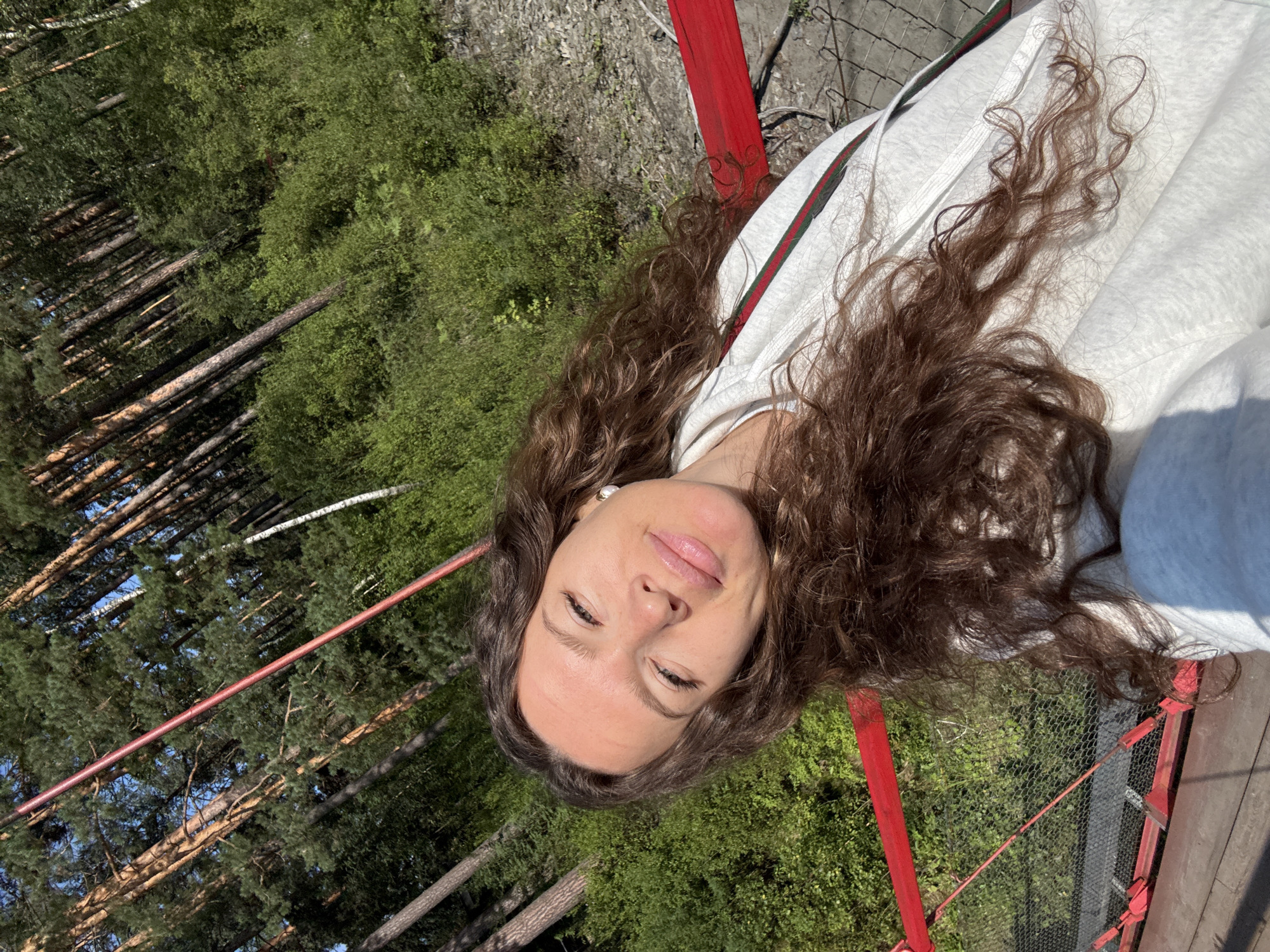 The image is a selfie taken by a young woman with long, curly brown hair, outdoors in a wooded area with a red railing in the foreground. The woman is wearing a light-colored hooded sweatshirt with a green and red stripe down the side. She is looking towards the camera with a slight smile. Sunlight is hitting her face. Behind her, a dense forest of pine trees and green foliage is visible under a clear blue sky. A red railing and a wire fence are part of the structure she is on, likely a bridge or elevated walkway. The location is described as being in the city of Barangol, Russia, suggesting a natural setting within or near the city.