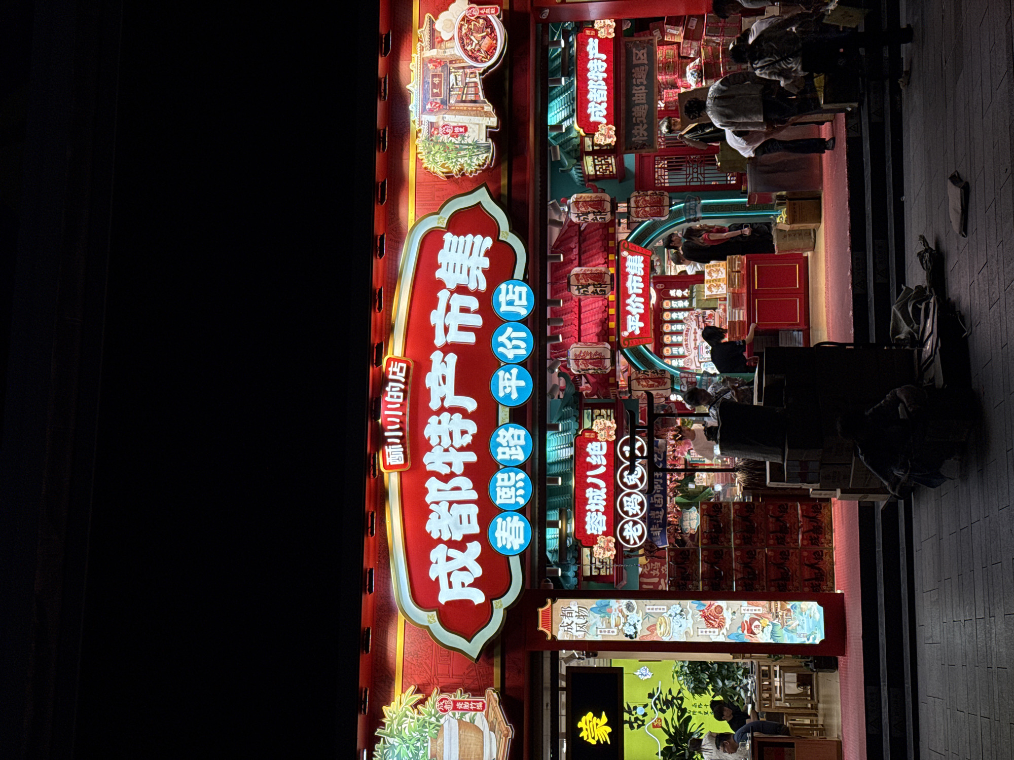 The image shows the facade of a store in Jinjiang District, China, likely at night given the darkness and illuminated signs. A vertical sign in red and white Chinese characters identifies the store as "Chengdu Specialty Market Collection". There are other illuminated signs with smaller Chinese characters, including one that reads "Suzhou Xiaoxiao Dian". The store's entrance area includes displays of products, some of which appear to be packaged in red boxes. People are visible inside the store and on the sidewalk outside. The lighting creates a vibrant and bustling atmosphere.