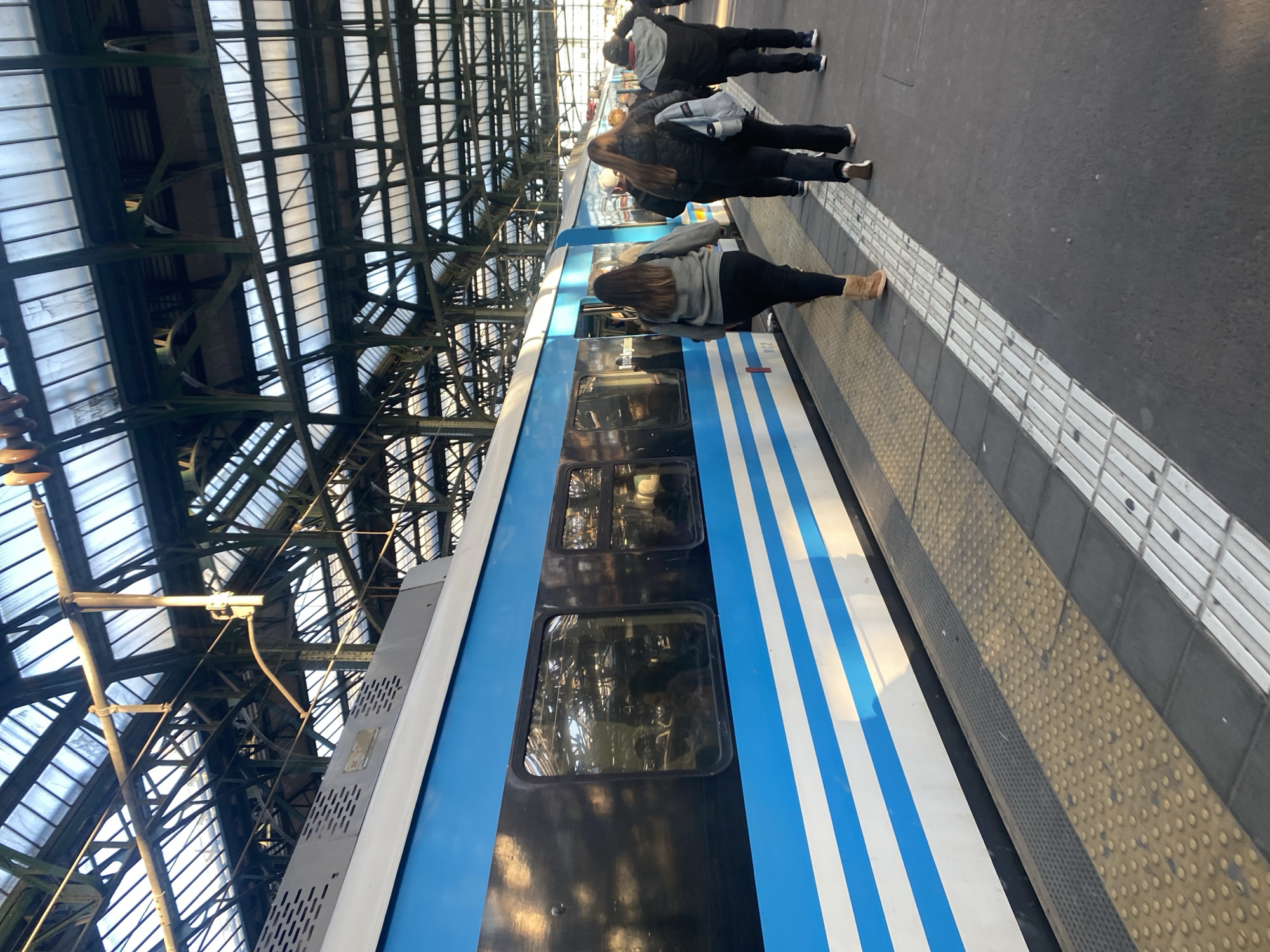 The image captures a train platform scene, likely in Buenos Aires, Argentina, given the distinct blue and white livery on the train car. Several people are standing near the train. The platform is paved with patterned tactile paving along the edge. The overhead structure of the station features a complex network of metal beams and supports. The train car has windows reflecting the platform and people.