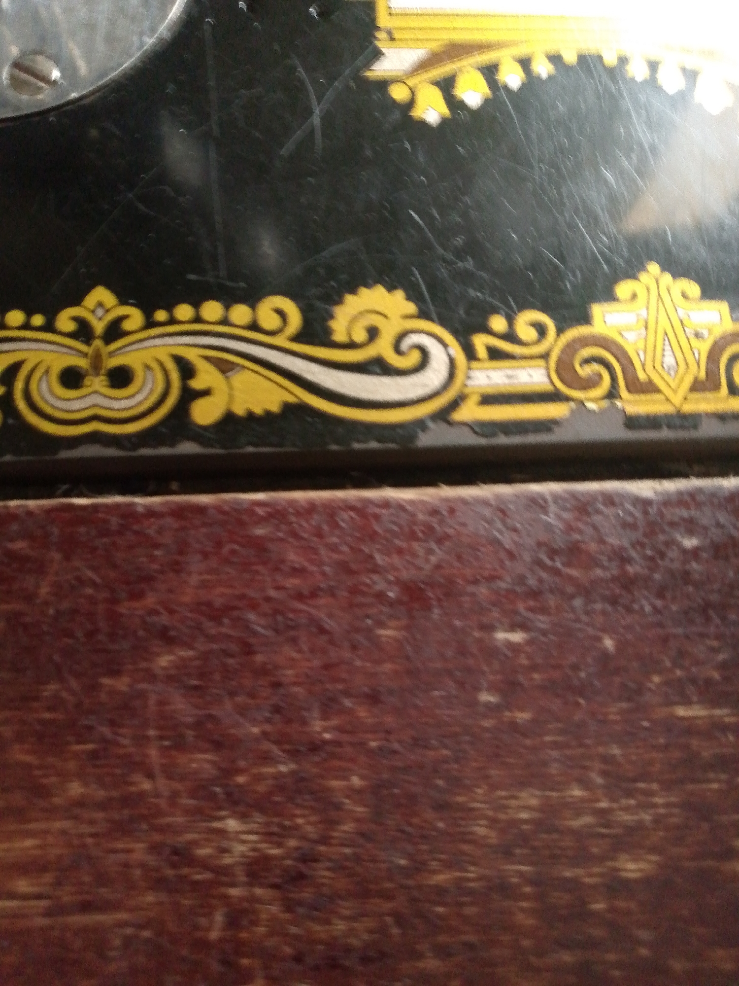 The image shows a close-up view of a dark, possibly antique, sewing machine. The top portion of the machine is black with ornate yellow and white decorative patterns. These patterns appear to be decals or painted designs, featuring swirls, dots, and geometric shapes. Part of a silver metal component with a screw head is visible in the upper left corner, likely part of the sewing machine's mechanism. Below the decorative band, a darker, possibly wooden surface is visible, characterized by a reddish-brown hue and a textured grain. This lower section appears worn, with visible scratches and variations in tone, suggesting age or use. The image is a still life, focusing on the details of the sewing machine's surface. There are no people or any indication of Jauro Yinu, Nigeria.