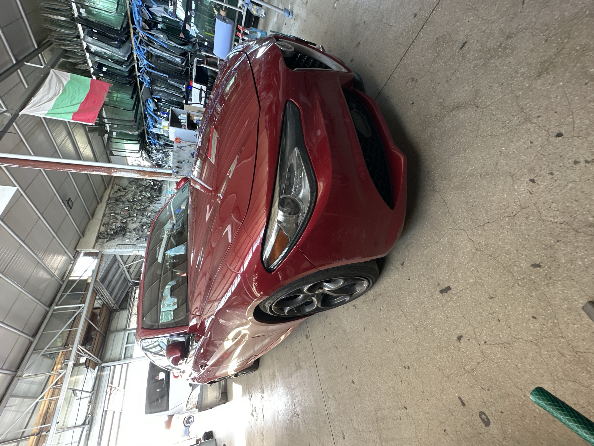 The image shows a red Alfa Romeo car parked inside a garage. The car is the primary subject, positioned in the foreground and taking up most of the frame. It is a low-angle shot, emphasizing the car's front and side. In the background, there are shelves filled with car windshields, suggesting the location is an auto repair shop or a car parts store. A Bulgarian flag is hanging from the ceiling. To the left of the Alfa Romeo, another white vehicle is partially visible. The floor is concrete and appears worn. Natural light is coming in from an unseen opening on the left side of the garage. The overall environment is industrial and functional.