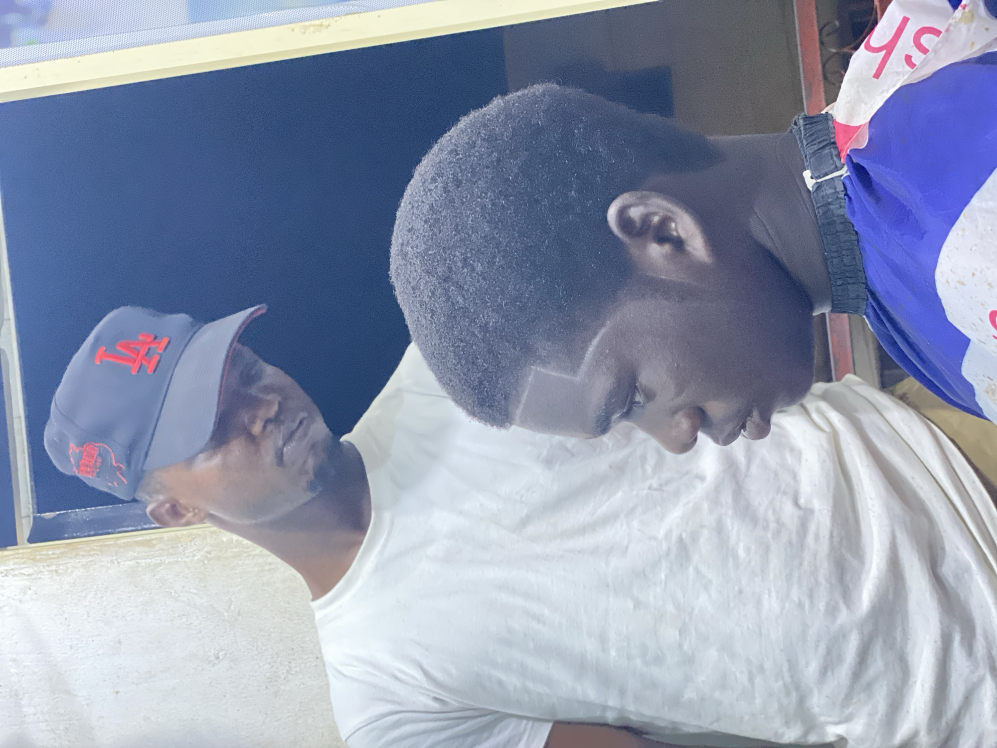 The photograph depicts two young men, one wearing a dark blue baseball cap with a red "LA" logo, the other with a close-cropped haircut featuring a shaved line detail. Both are wearing white t-shirts. The environment is enclosed, possibly indoors. The city is specified as Ado, Nigeria.