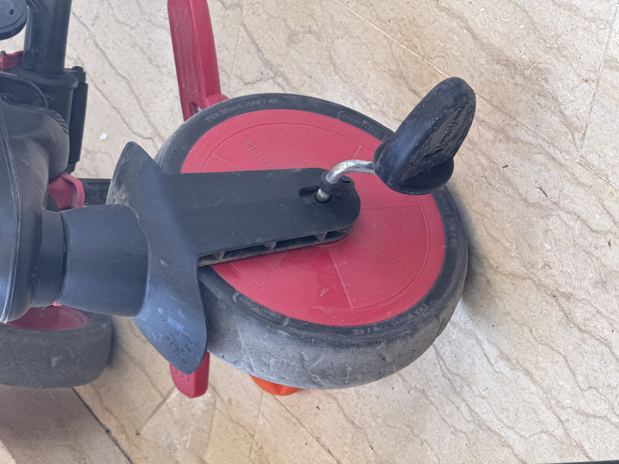 This media file is a close-up, overhead shot of a red and black tricycle, likely a child's toy, resting on a tiled floor. The tricycle's features, including its red wheel with a black tire and a black plastic guard, are prominently displayed. The wheel has "EVA Wheel 220/40" and "Smartrike" embossed on its side. The surrounding floor is tiled with a light beige, marbled pattern, with some visible grout lines indicating it's an indoor or covered outdoor space. The lighting suggests it is daytime, with no strong shadows indicating a specific time of day, and the overall scene is neutral in terms of weather. The tricycle appears to be stationary, with no people or active event taking place. The focus is on the object itself and its immediate surroundings. The location context provided is Boreti, Montenegro, but there are no specific visual cues within the image to confirm this.