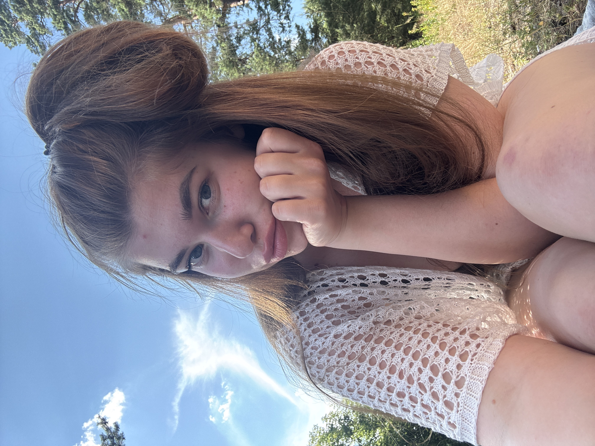The media file shows a young woman with long, auburn hair styled in a bun on top of her head. She is lying down outdoors, with her head resting on her hand. The woman is wearing a white, crocheted top. The background consists of a blue sky with some clouds, trees, and dry vegetation. The lighting suggests it is daytime, likely with bright sunlight. The woman's expression appears relaxed or pensive. There is no visible text in the image. The provided location context is Kurovskoye, Russia.