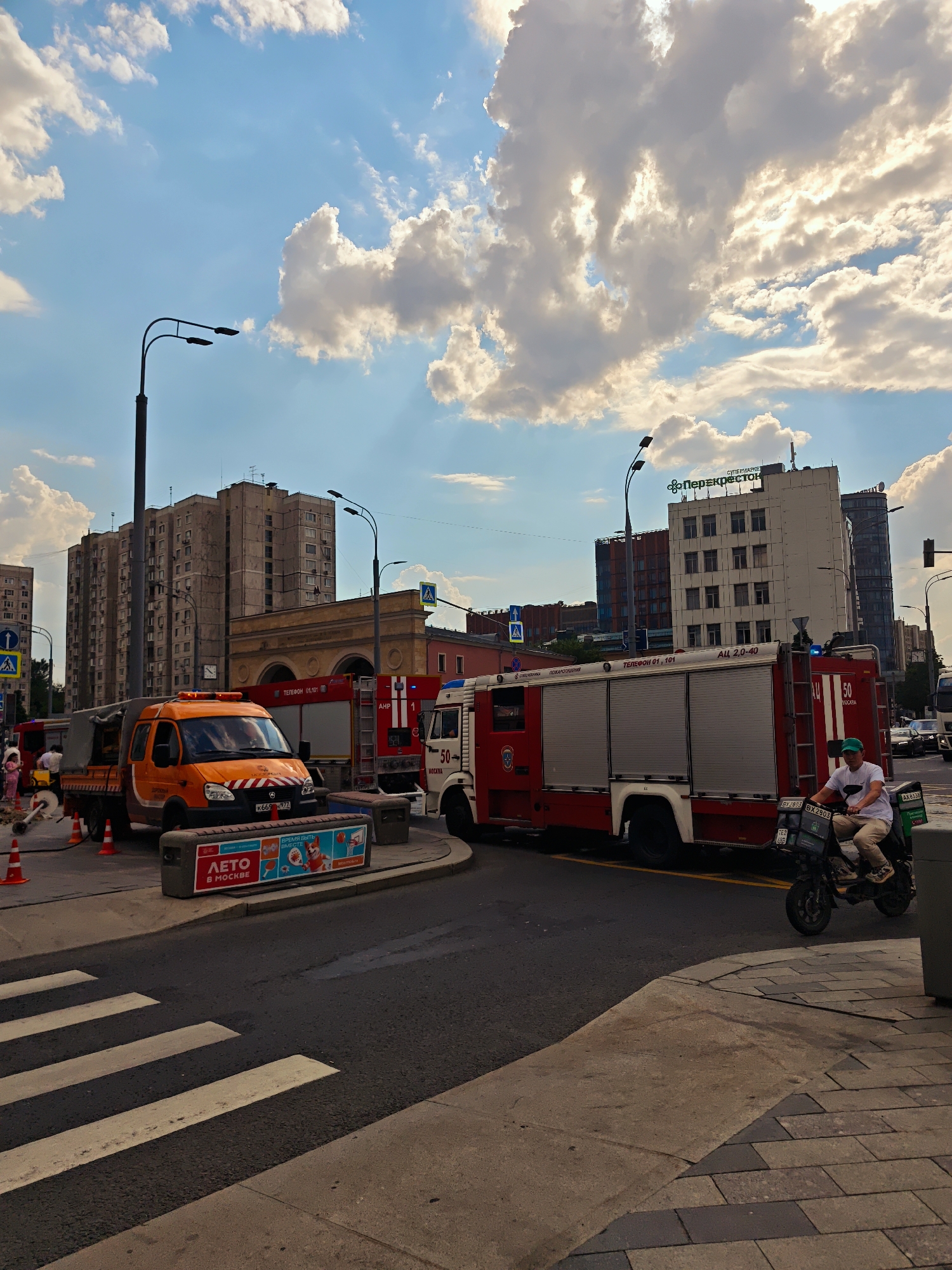 In an urban setting under a partially cloudy sky, a street scene unfolds with a focus on emergency vehicles and urban infrastructure. Two fire trucks, one predominantly red with white trim and the other partially visible in orange, are parked on the road. The red fire truck has "50" and "ПОЖАРНАЯ" (POZHARNAYA) visible on its side. An orange utility truck with a covered bed is positioned to the left, with several people standing near it, appearing to be involved in some activity or operation. Orange traffic cones are placed on the road near the utility truck, suggesting a work zone or temporary obstruction.
A man wearing a green cap, white t-shirt, and light-colored pants is riding a dark-colored scooter with green delivery boxes attached to the back. He is moving from right to left in front of the red fire truck. The scooter has license plates visible, including "AX 835X".
The background reveals tall, multi-story buildings, characteristic of a city environment. One building is lighter in color with a sign at the top that reads "ПЕРЕКРЕСТОК" (Perekrestok). Other buildings are darker and appear residential or office spaces. Streetlights and traffic signals are also visible.
In the foreground, a pedestrian crossing with white stripes on asphalt is prominent. The ground texture shifts from asphalt to a paved sidewalk area composed of rectangular stones. A low, gray concrete barrier with a colorful advertisement featuring text like "ЛЕТО В МОСКВЕ" (Summer in Moscow) and images of children and a dog is situated between the road and the sidewalk. The overall impression is one of a busy city intersection with ongoing operations.