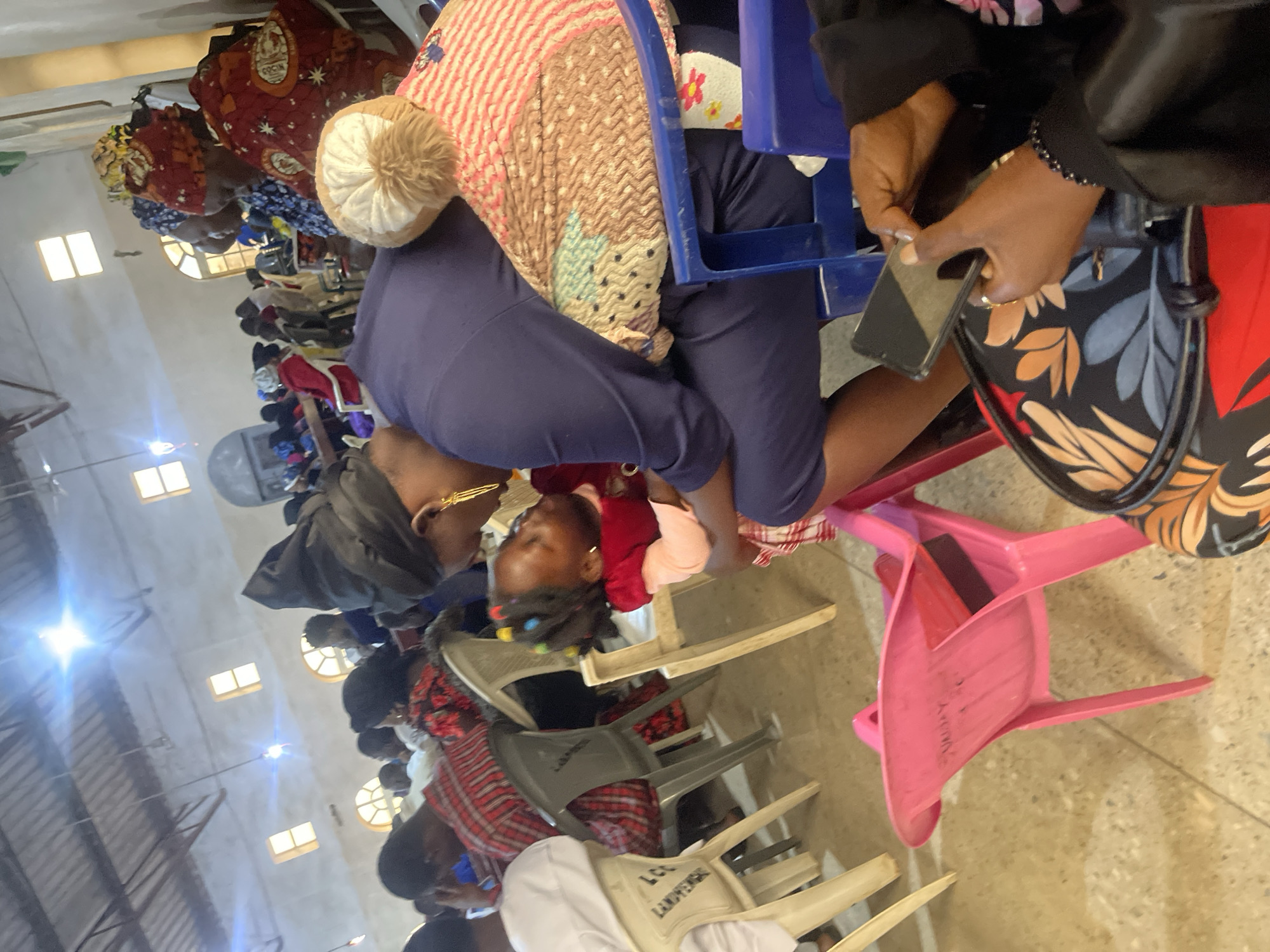 The image depicts an indoor gathering, possibly a church or community hall, in Jos, Nigeria. In the foreground, a woman in a dark blue outfit is embracing a young child. The woman is wearing a black headscarf and large gold earrings. The child, with colorful beads in their hair, is dressed in a red and light pink outfit and a checkered skirt. They are positioned in front of a pink plastic chair. To the right, another person's hands are visible holding a smartphone. In the background, several other individuals are seated, some wearing colorful attire, suggesting a festive occasion. The environment appears functional, with a corrugated metal ceiling and fluorescent lighting. The seating arrangement consists of plastic chairs of various colors, including white and gray. The overall scene suggests a moment of connection and community within a public indoor space in Jos.