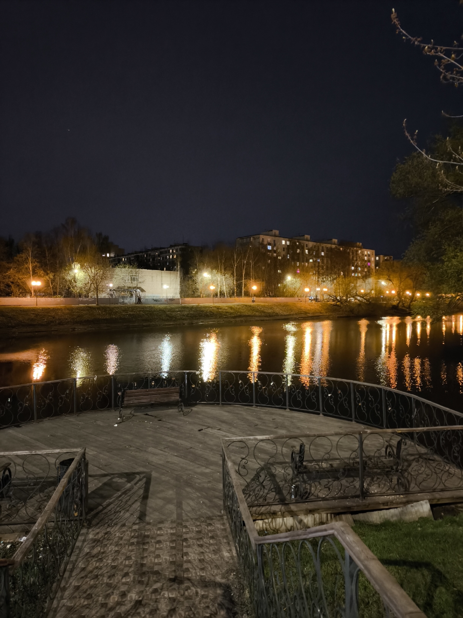 The image presents a serene nighttime view of a lake or pond in Khimki, Russia, observed from a wooden viewing platform. In the immediate foreground, a dark wooden platform with ornate, dark metal railings extends into the scene. Two park benches with wooden slats and metal frames are situated on the platform, facing the water. Shadows, possibly from overhead lighting, are cast across the platform's textured surface.
Across the water, the opposite bank is lined with leafless trees, illuminated by a series of warm-toned streetlights. These lights create striking, elongated reflections that shimmer vibrantly on the water's surface, ranging in color from bright white to orange. Behind the treeline and streetlights, several multi-story buildings are visible. On the right, a large apartment building with numerous lit windows stands prominently, indicating residential activity. On the left, a lighter-colored building or wall is present, near which a small gazebo-like structure can be seen. A solitary figure appears to be walking on the far bank near the gazebo.
The sky above is dark and clear, with a single faint, bright point of light visible in the upper left, suggesting a star or a distant aerial object. A bare tree branch with small buds extends into the upper right corner of the frame, silhouetted against the night sky. The scene is illuminated entirely by artificial light sources, creating a tranquil and atmospheric setting. No discernible text is visible within the image.