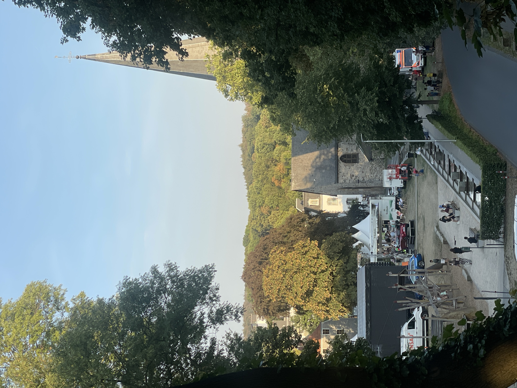 The image depicts an outdoor event or festival taking place in a town square, likely in the vicinity of a church. In the foreground, on the left, is a large tree with green foliage. In the middle ground, there's a black stage structure, a white van with "REMSAU" written on it, and a blue car parked nearby. Further back, several white tents are set up, suggesting vendor stalls or event attractions. People are visible milling around the tents and sitting on benches. To the right of the tents is a stone building, possibly part of a church, with a tall, pointed steeple topped with a cross reaching into the clear blue sky. The background consists of a dense forest with trees displaying a mix of green and autumnal colors. On the right side of the image, a paved road curves away, with more trees lining the roadside. An ambulance is parked near a group of people. The overall scene suggests a community gathering in a semi-urban, wooded environment. This scene is consistent with the area around the Evangelische Kirche Menden-Lendringsen or a similar church in Iserlohn, Germany, which often hosts community events.